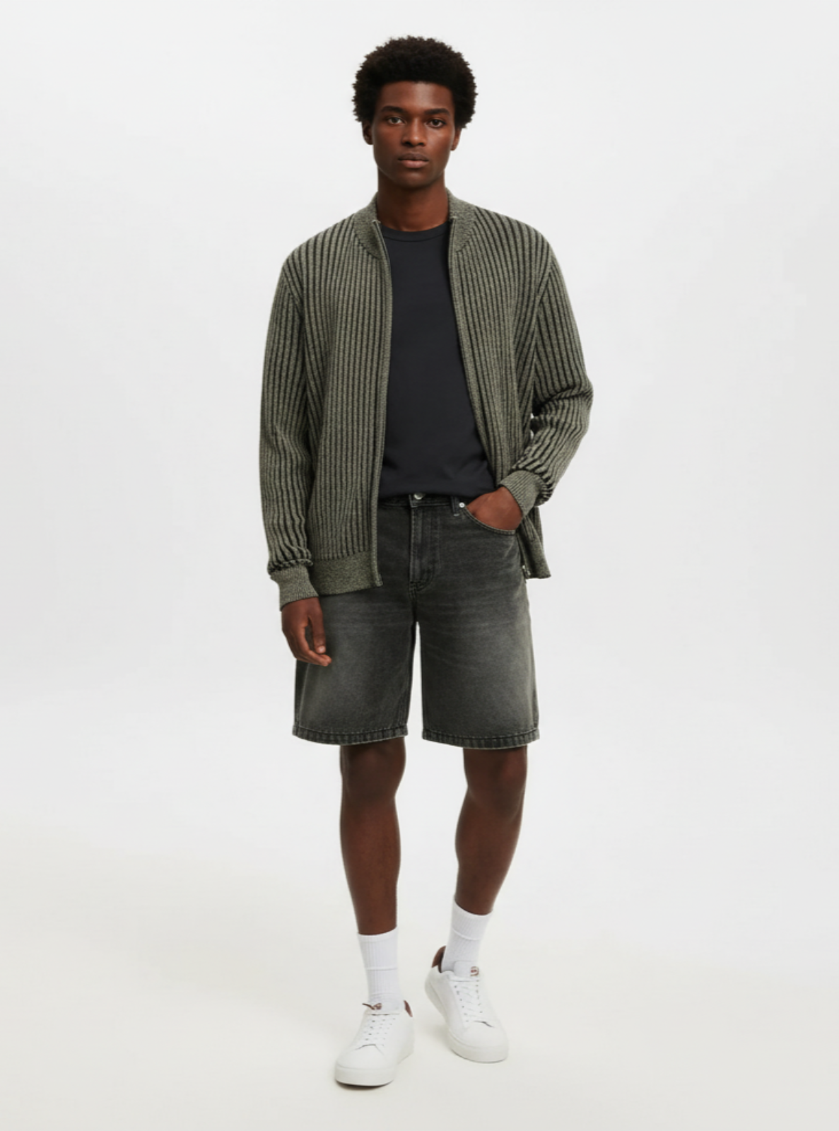 Man wearing a green cardigan, black shirt, gray shorts, and white sneakers on a white background