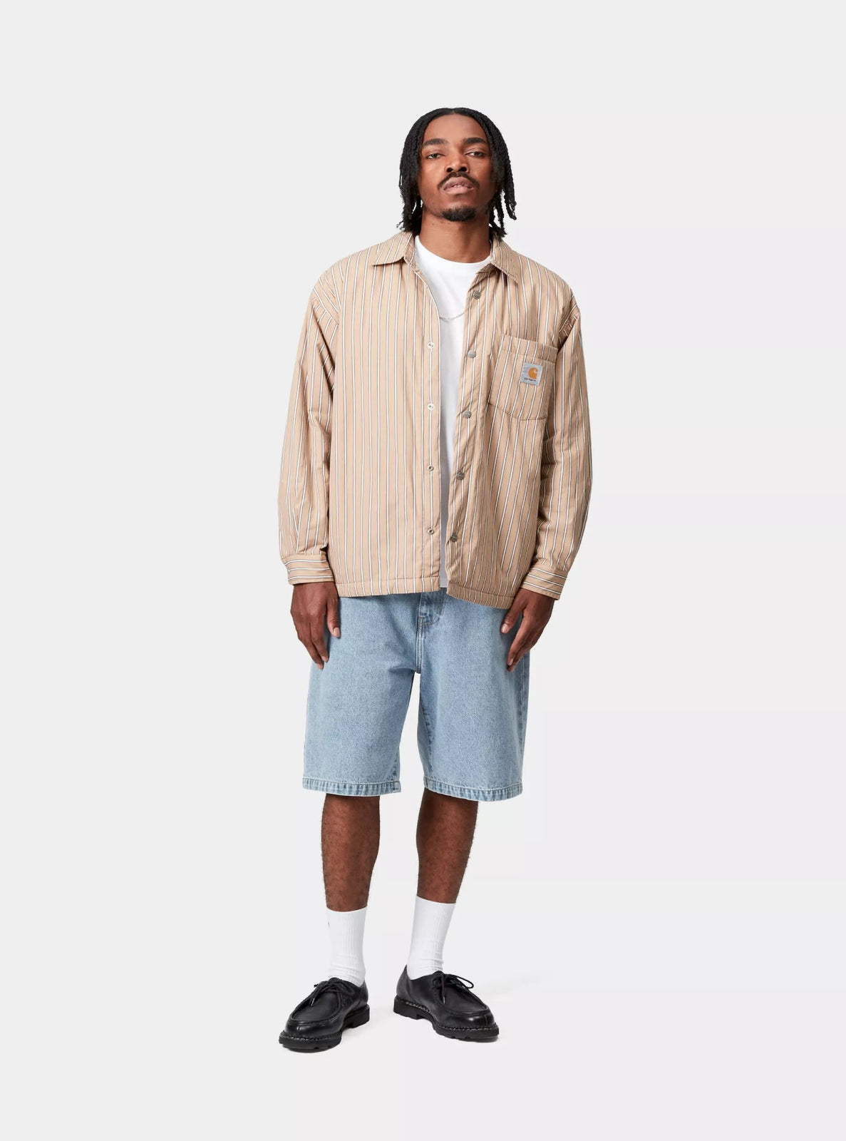 Man wearing a beige jacket, white shirt, light blue shorts, and black shoes on a white background