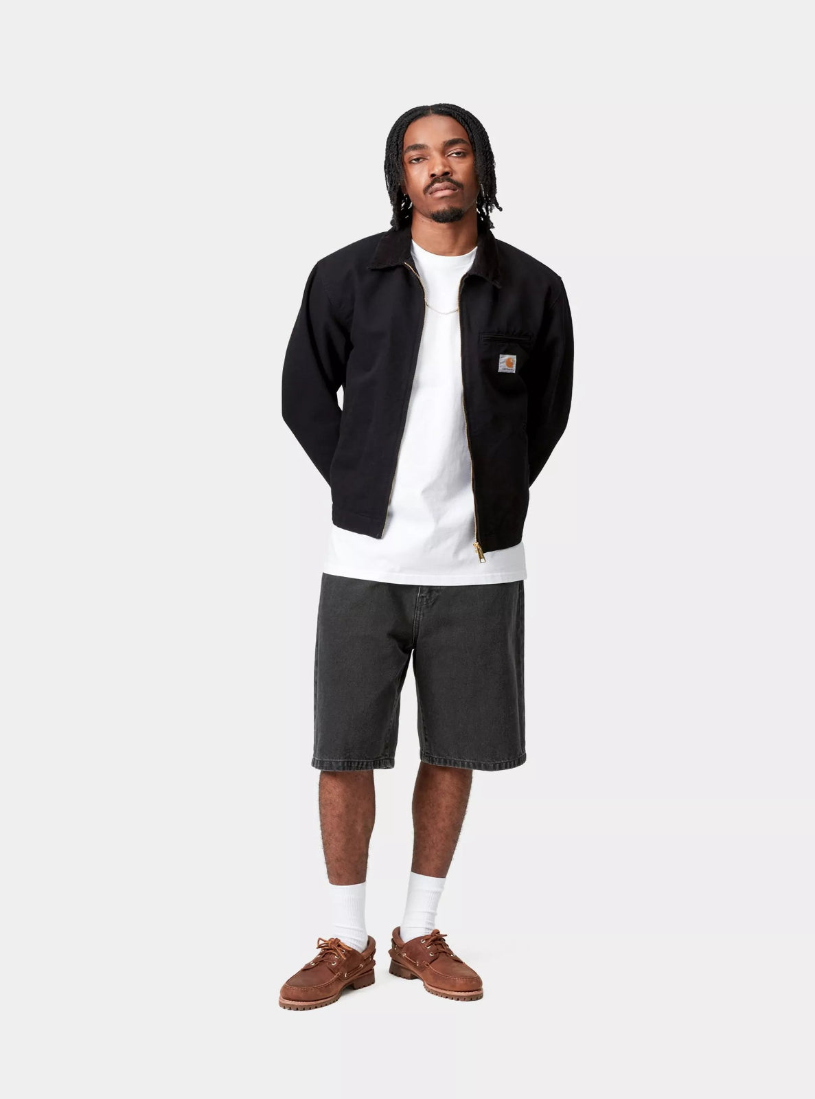 Man wearing a black jacket, white t-shirt, gray shorts, and brown shoes on a white background