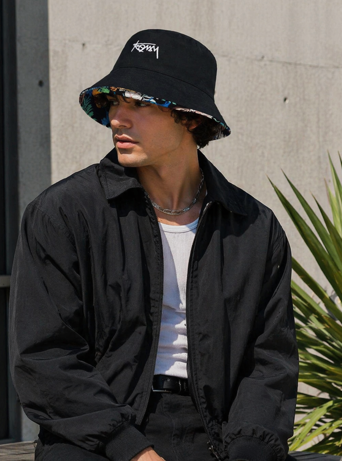 Person wearing a black bucket hat with a brand logo, black jacket, and white shirt against a concrete wall.