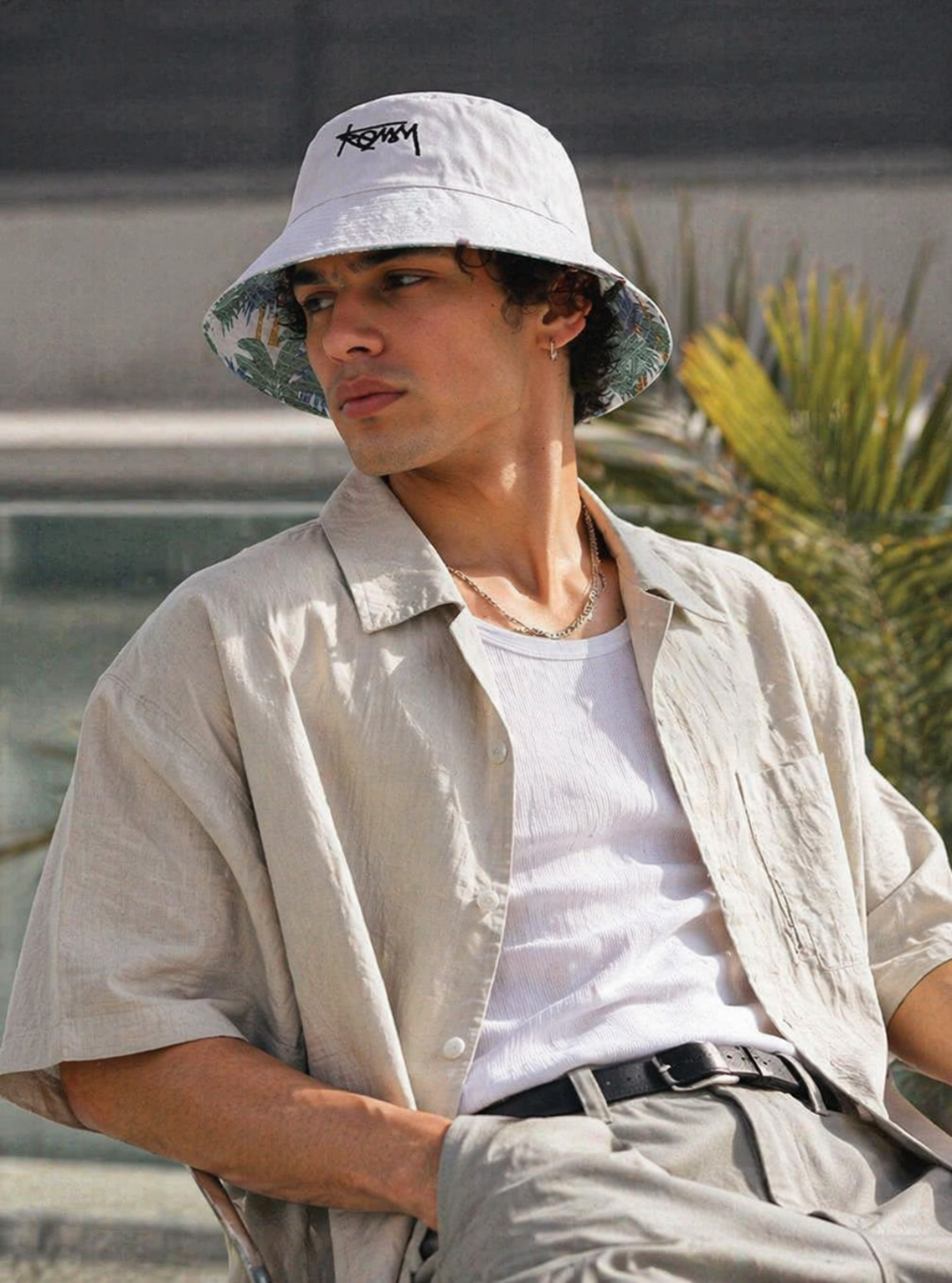 Man wearing a white bucket hat with a brand logo, sitting outdoors.