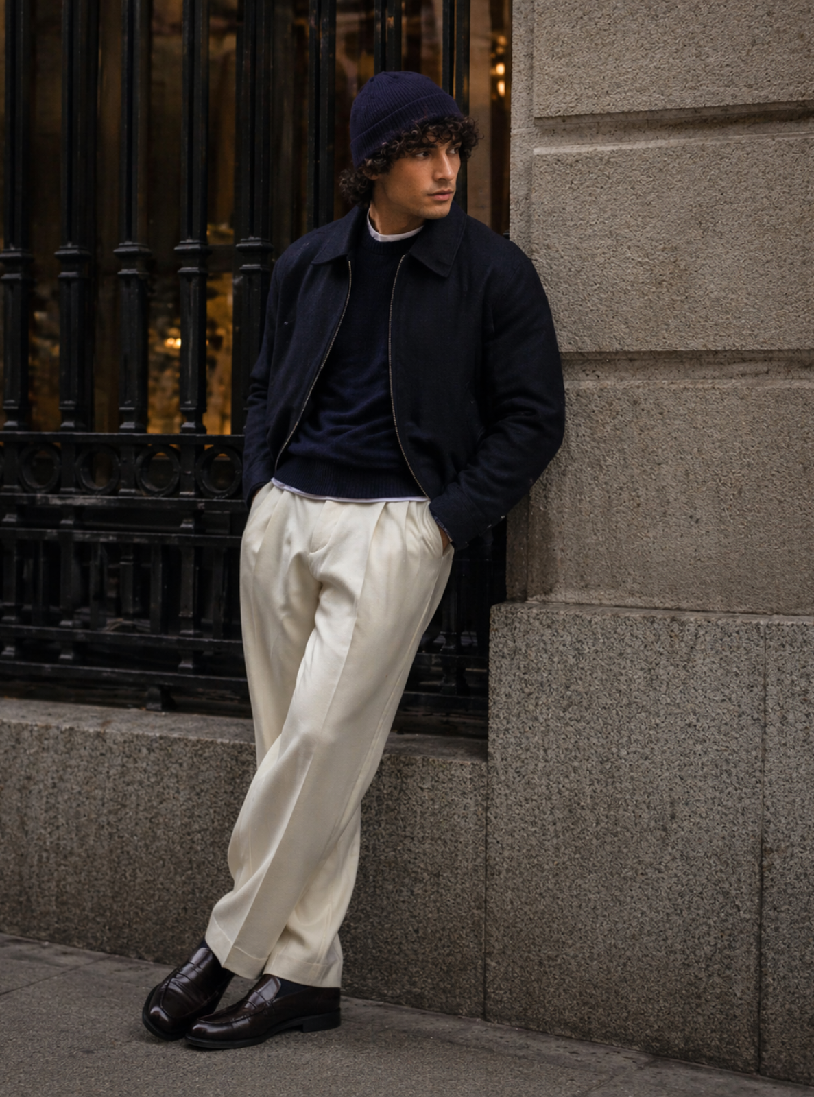 Person wearing a dark jacket, white pants, and black shoes leaning against a stone wall.