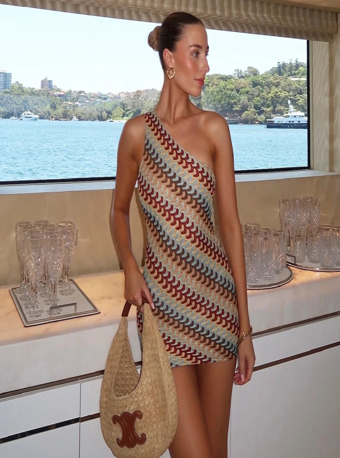Woman in a colorful one-shoulder dress holding a straw bag on a boat with water and skyline in the background.