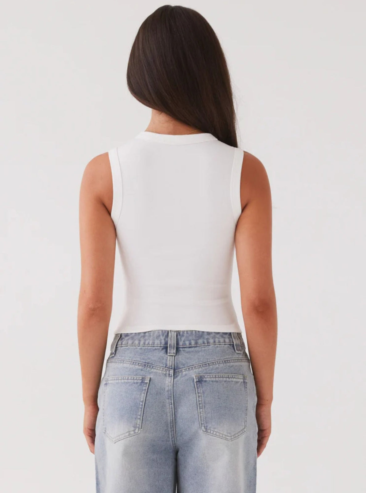 Person wearing a white sleeveless top and light blue jeans on a plain background