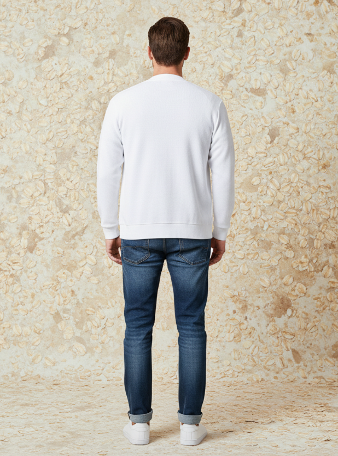 Man wearing a white sweater and blue jeans standing against a textured beige wall.