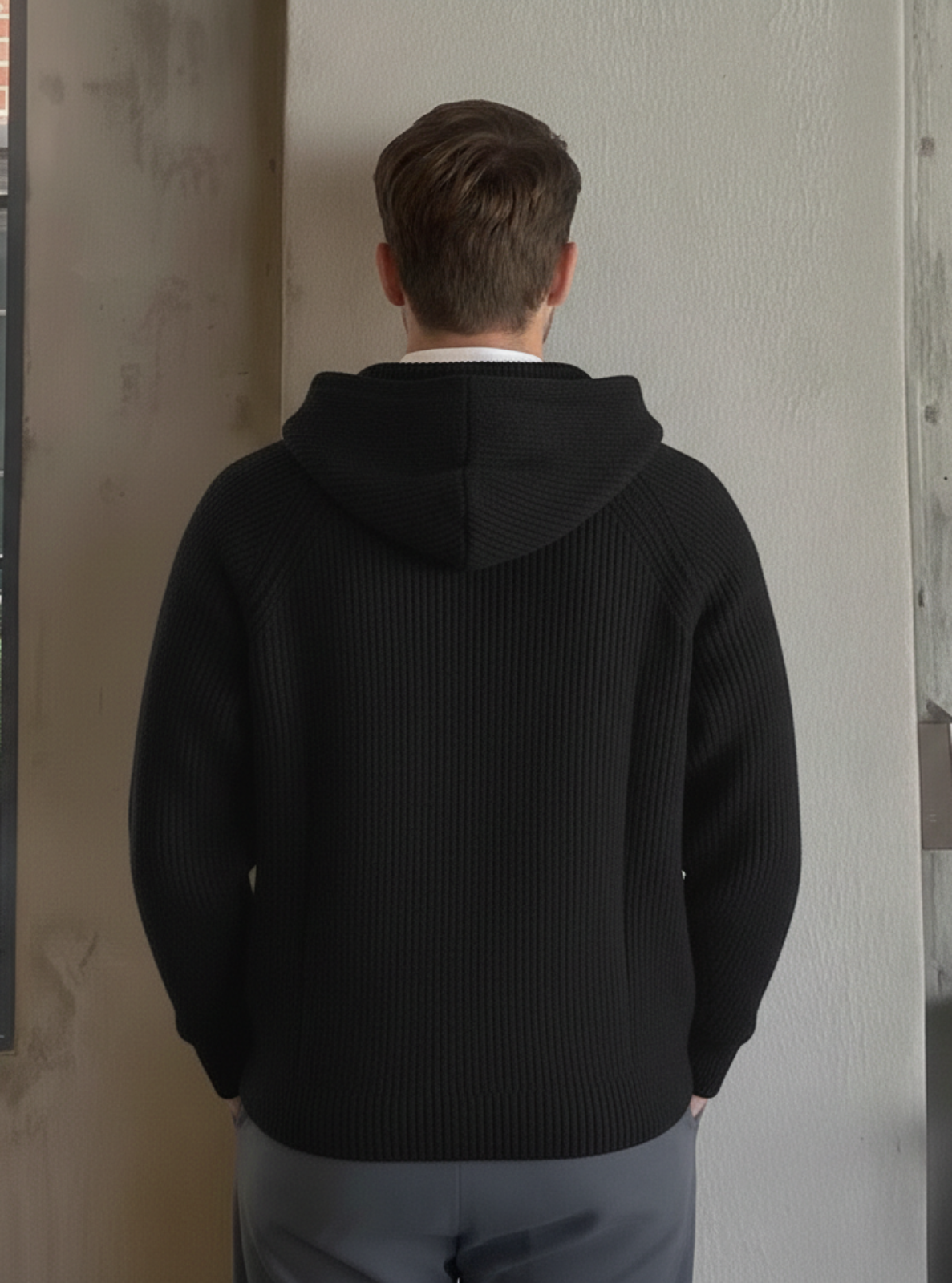 Person wearing a black hoodie standing in a room with a window and concrete wall.