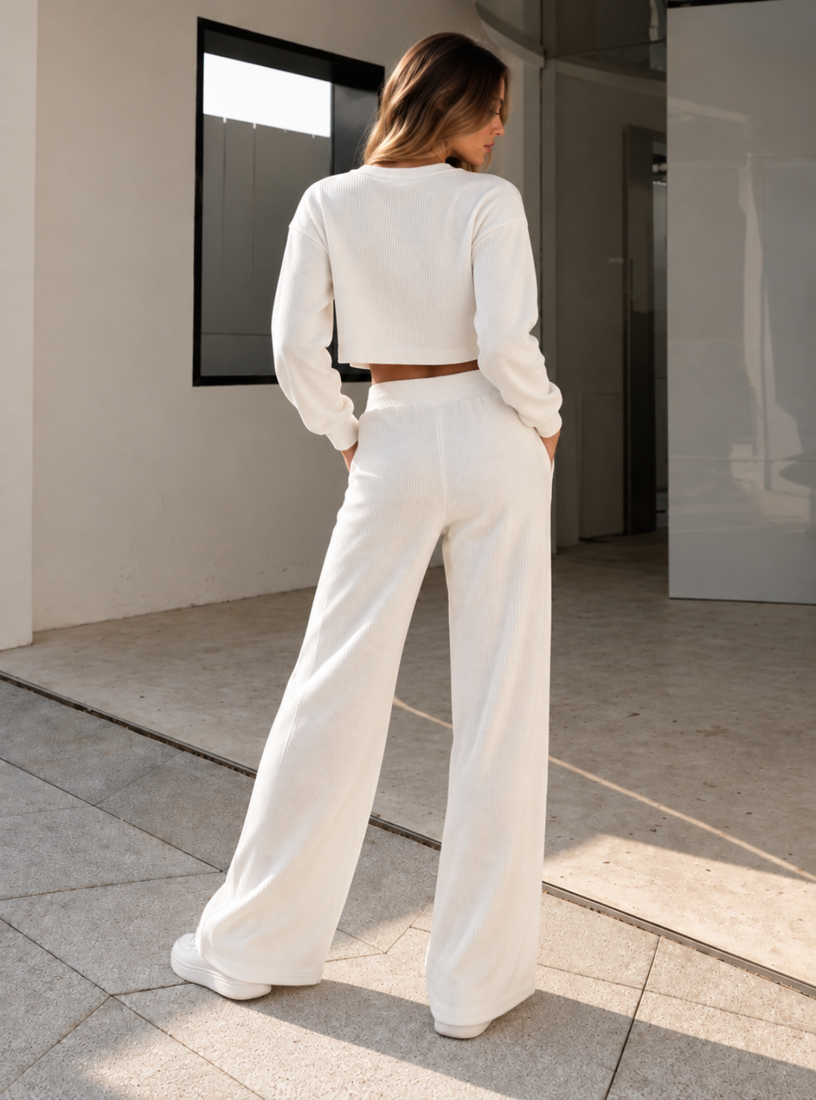 A woman stands with her back to the camera wearing a matching white long-sleeve crop top and wide-leg pants set in a modern indoor-outdoor architectural setting with natural light.