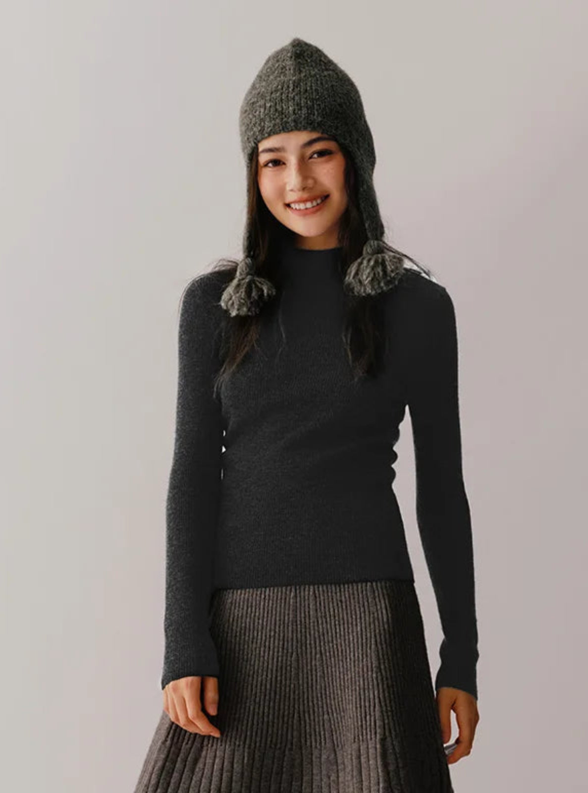 Woman wearing a dark sweater and knitted hat with pom-poms against a plain background