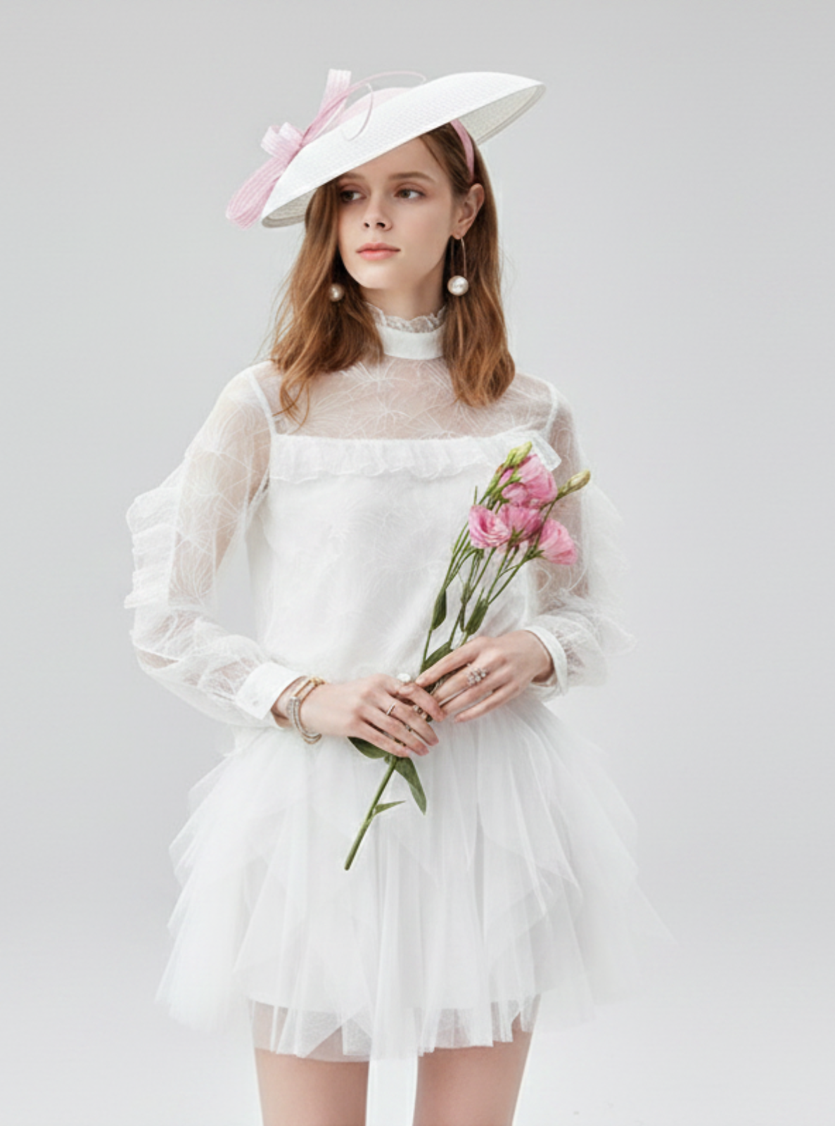 Woman in a white dress with long sleeves and a hat holding flowers on a light gray background