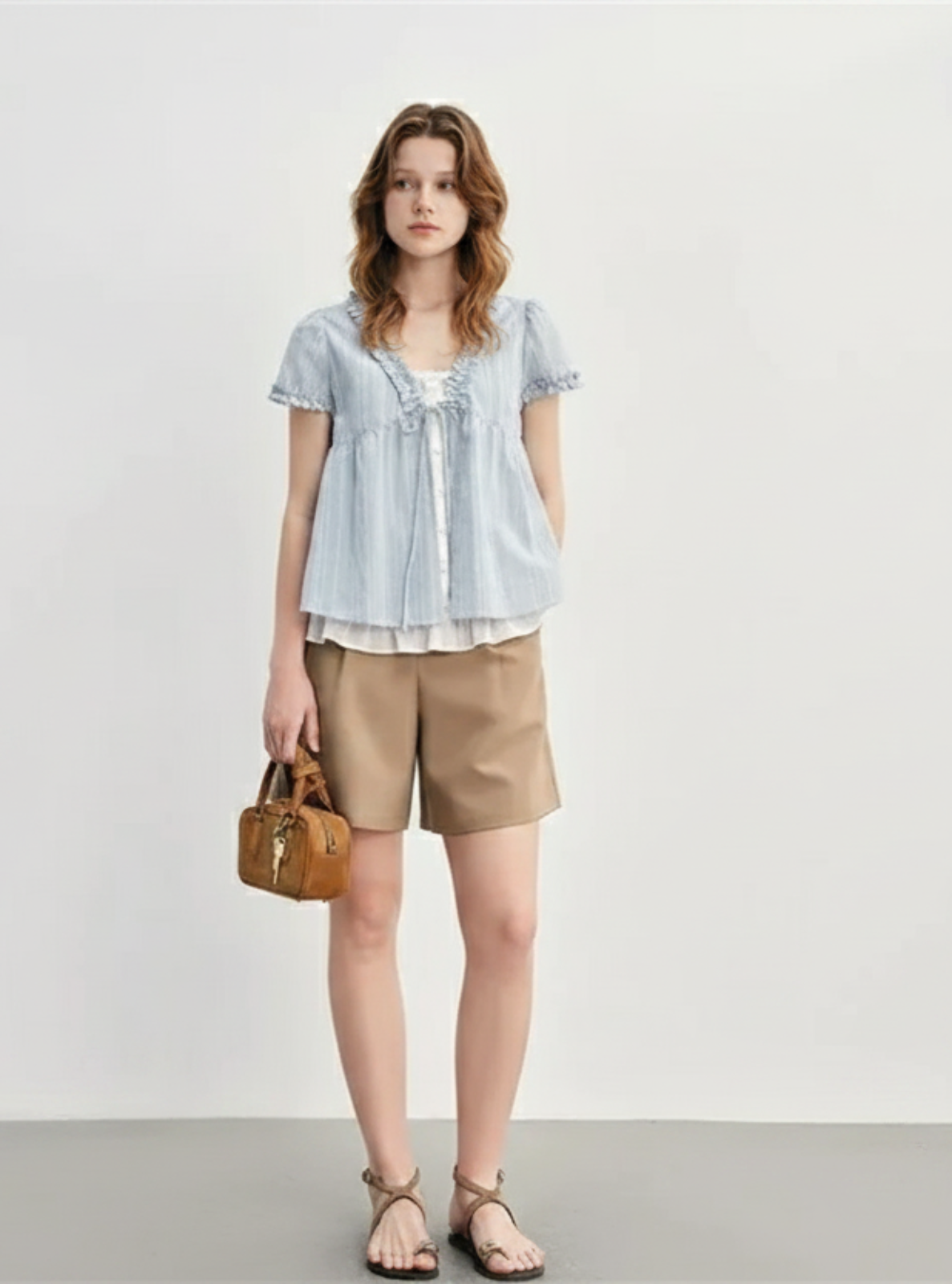 Woman wearing a light blue striped shirt and beige shorts, holding a brown handbag against a white background.