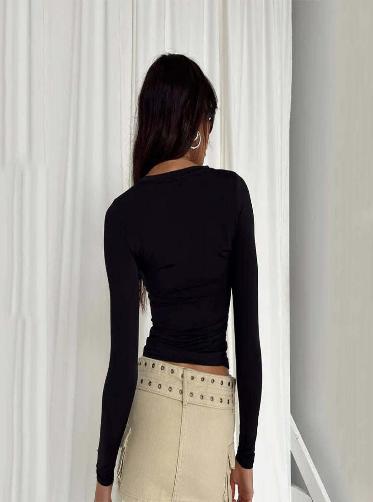 Person wearing a black long-sleeve top and beige skirt with white curtains in the background