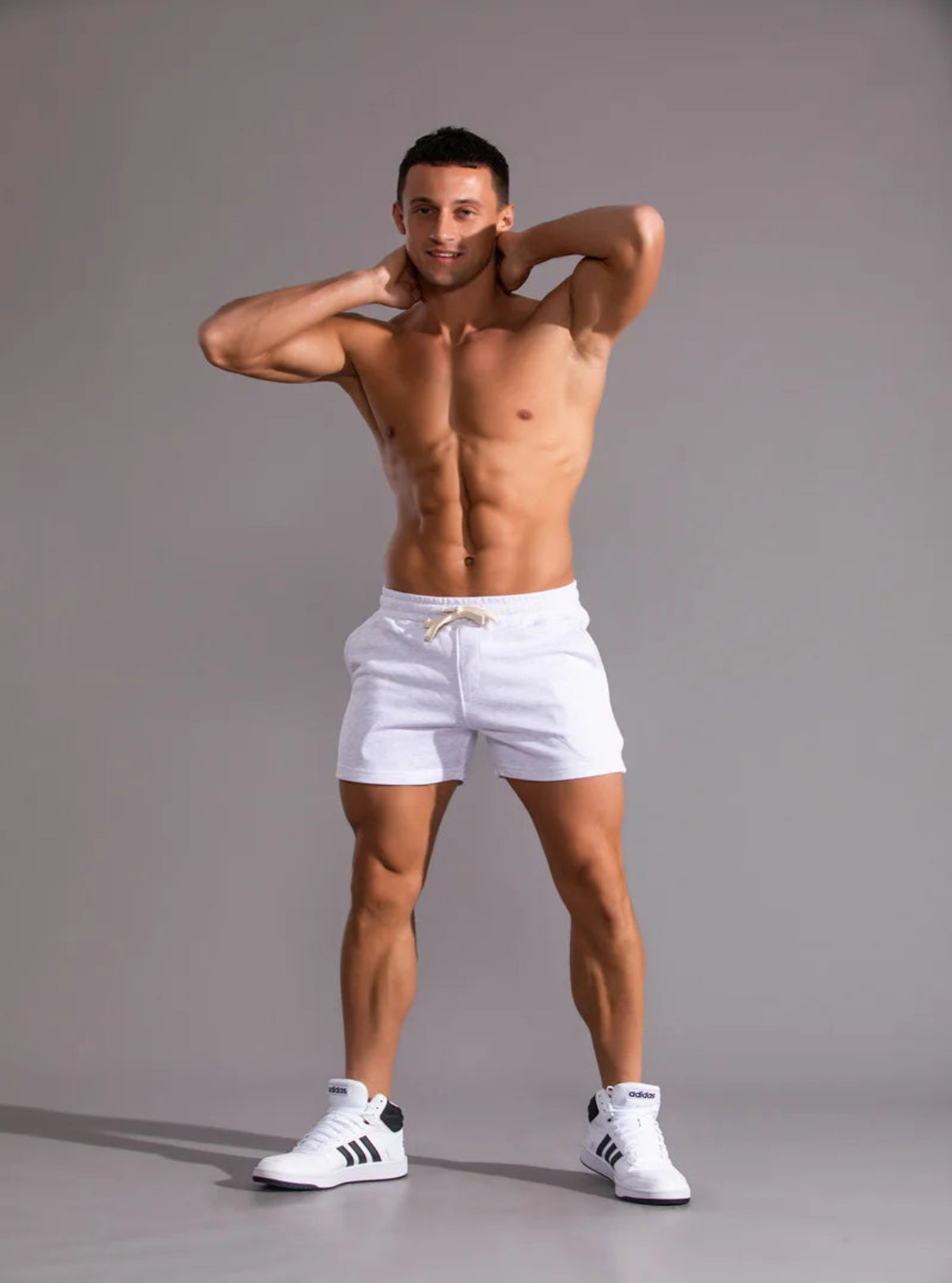 Muscular man wearing white shorts and black Adidas shoes on a gray background