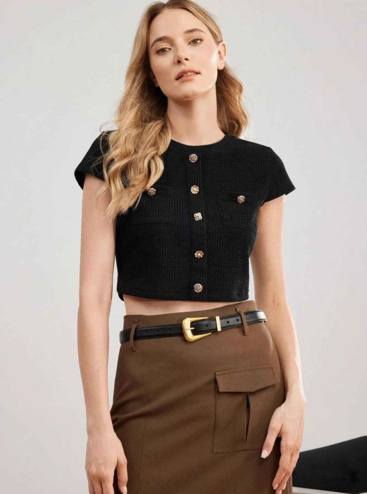 Woman wearing a black cropped top with gold buttons and brown pants with black belt on a light gray background