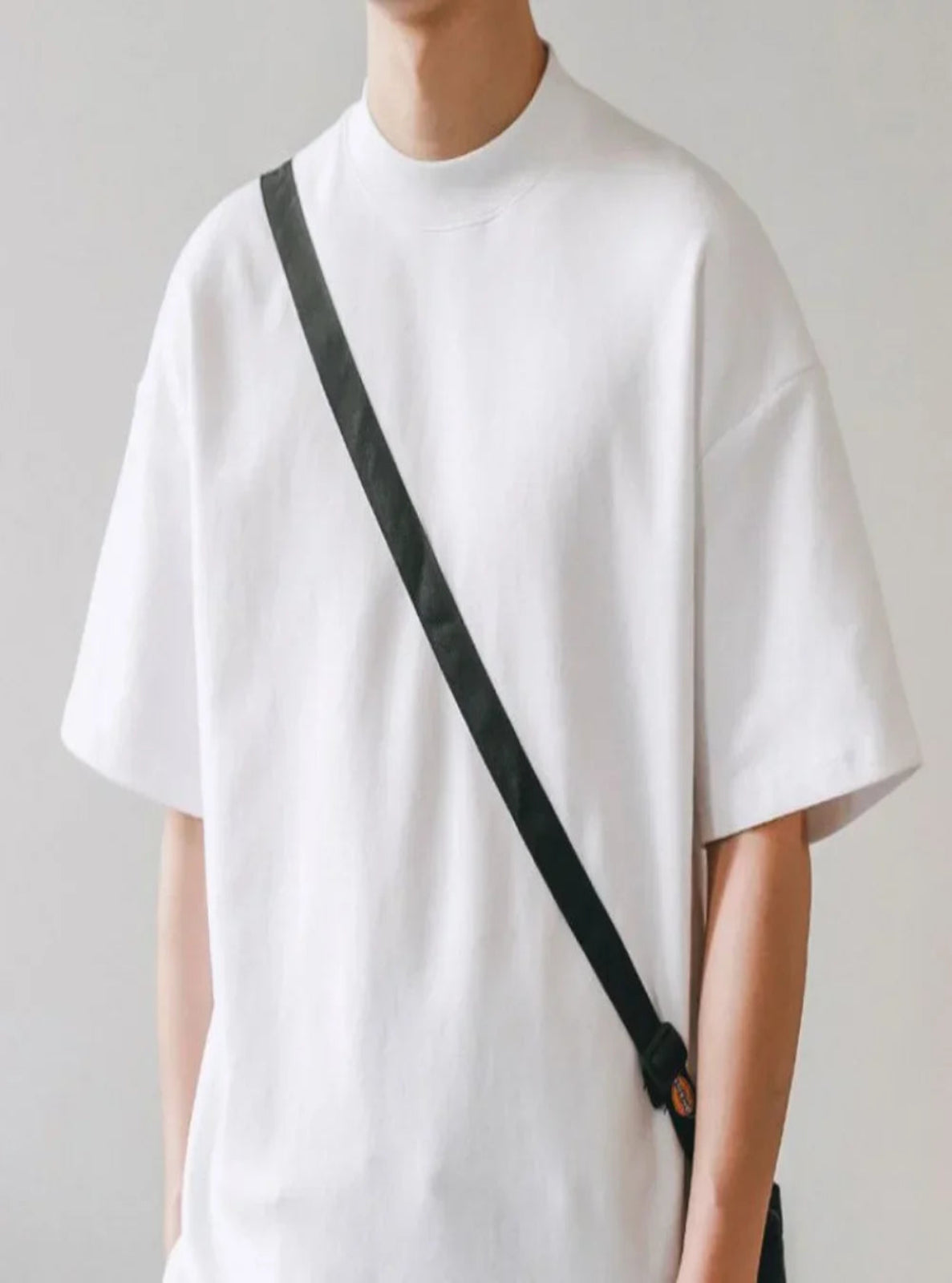 Person wearing a plain white t-shirt with a black strap over one shoulder against a light gray background