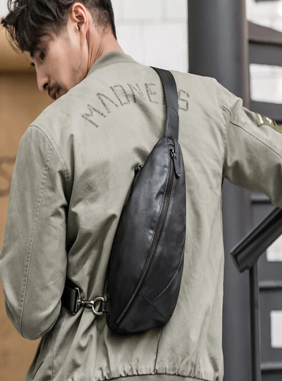Man wearing a green jacket with a black crossbody bag against a white tiled wall.