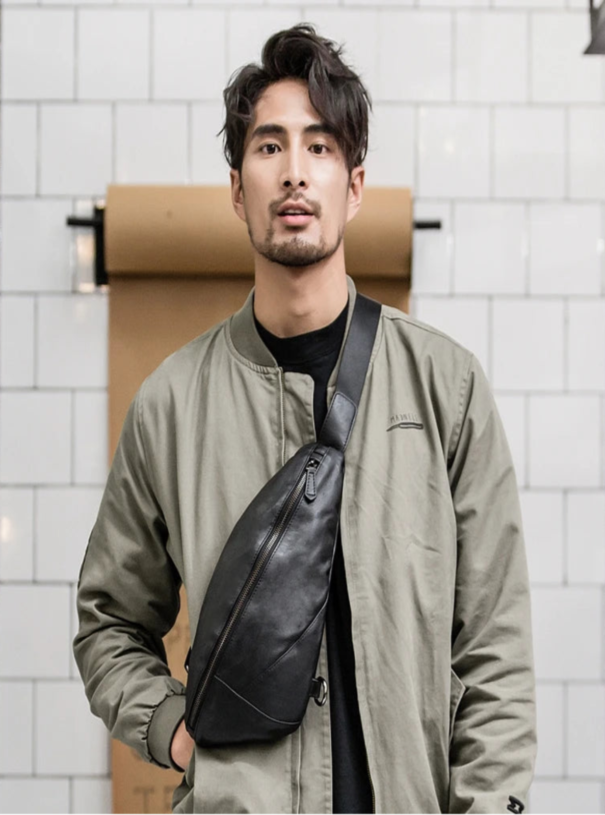 Man wearing a green jacket with a black crossbody bag against a white tiled wall.