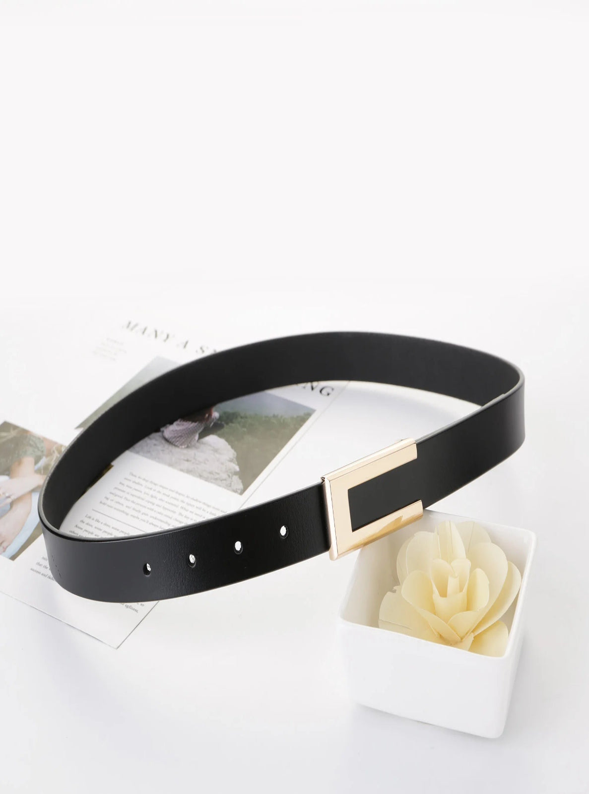 Black belt with a gold buckle on a white surface with a flower and magazine.