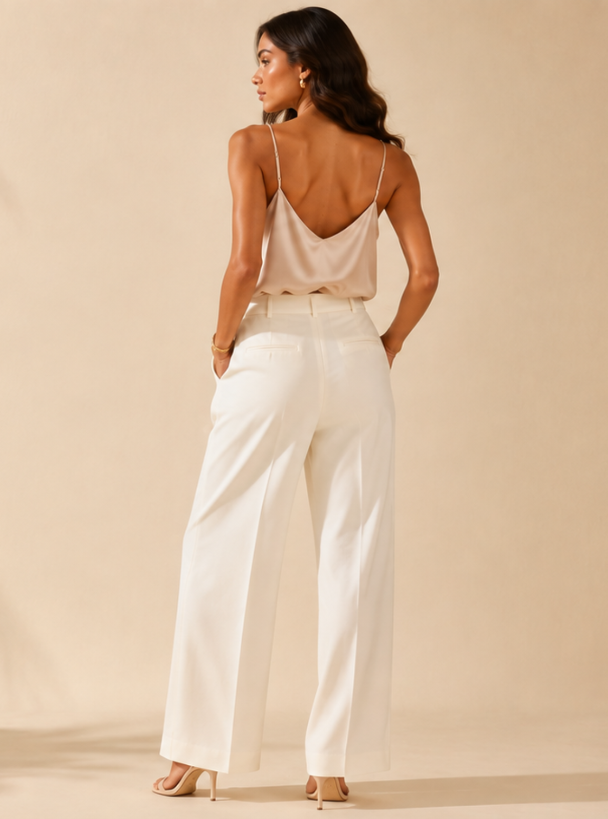 A woman stands with her back to the camera wearing a white satin camisole with a low open back and matching white wide-leg trousers, set against a warm beige background with palm leaf shadows.