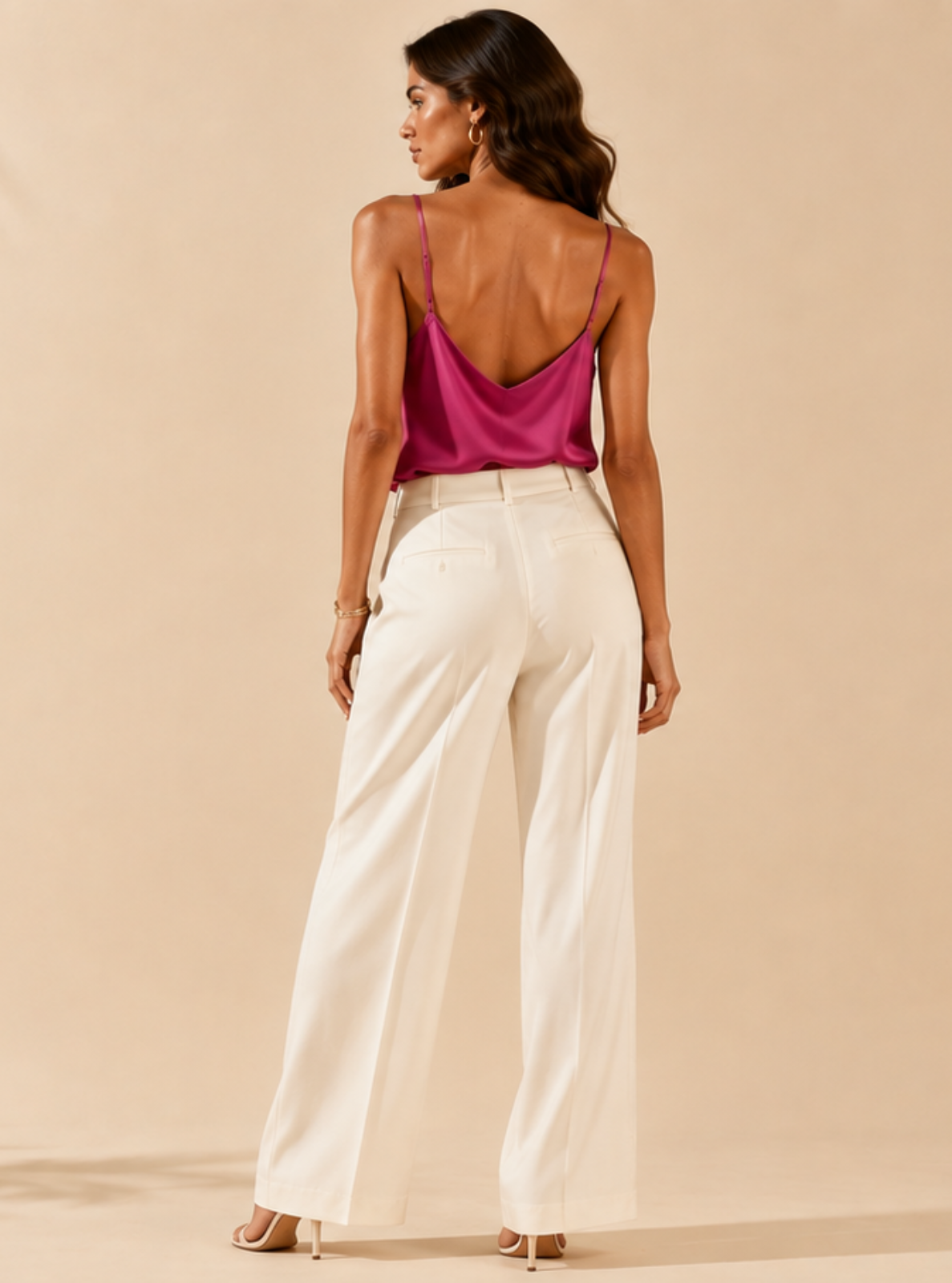 A woman stands with her back to the camera wearing a bright magenta open-back camisole and high-waisted white wide-leg trousers, set against a warm beige backdrop with palm leaf shadows.