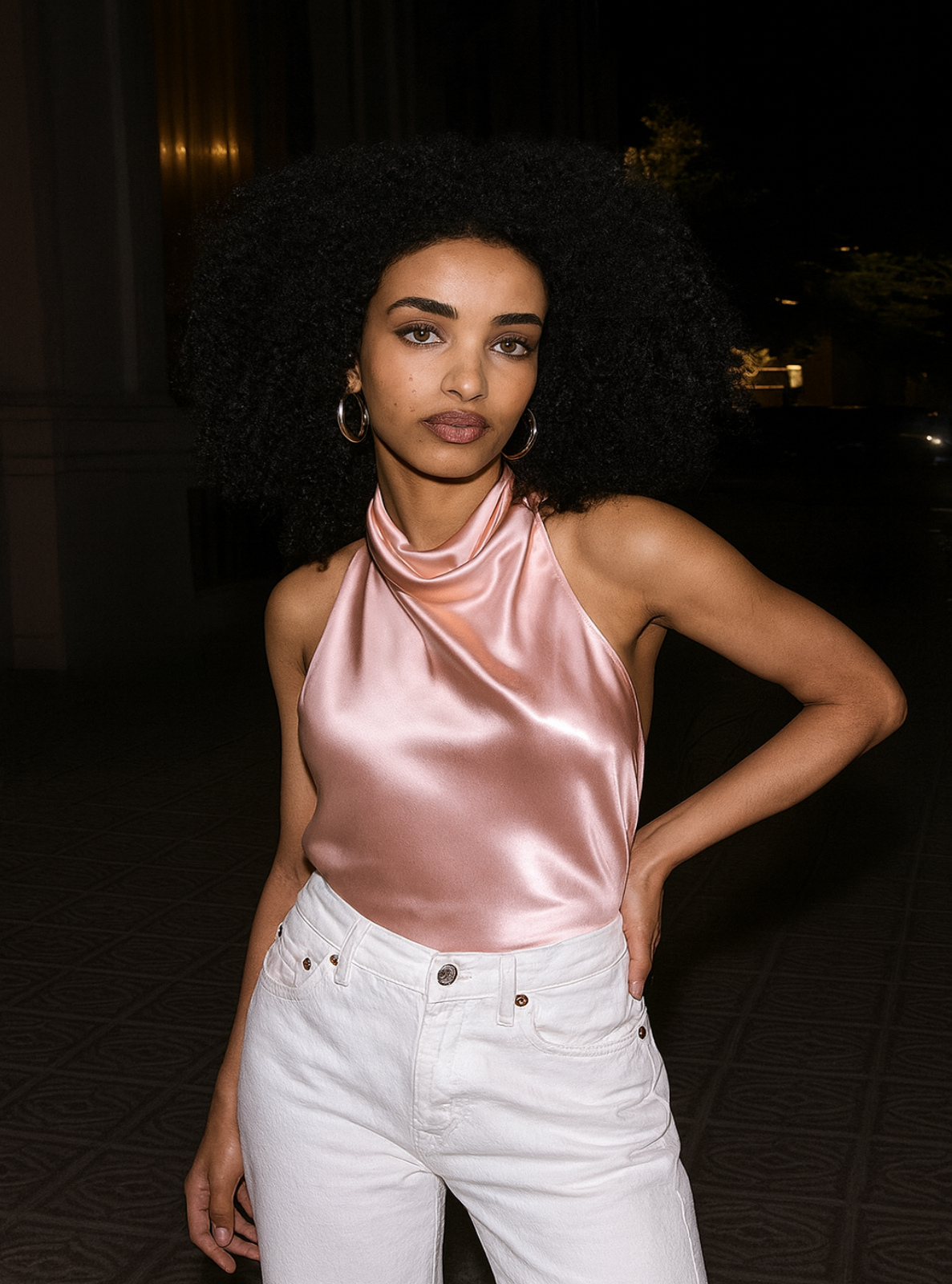 Woman wearing a pink satin top and white pants in a dark setting