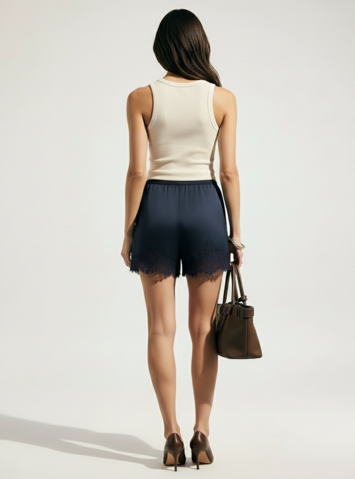 Woman wearing a beige sleeveless top and dark shorts, holding a brown handbag on a white background