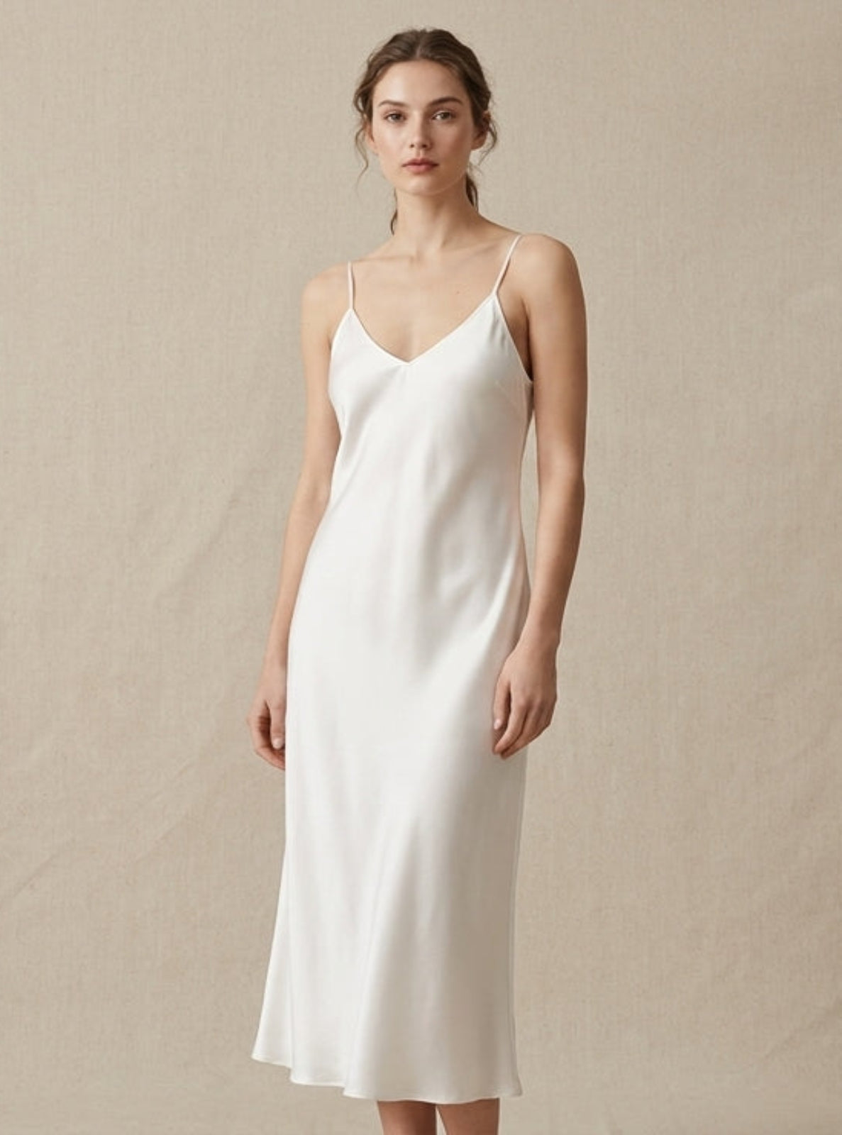 A woman modeling a white satin slip dress with thin spaghetti straps and a V-neckline, standing against a neutral beige textured backdrop.