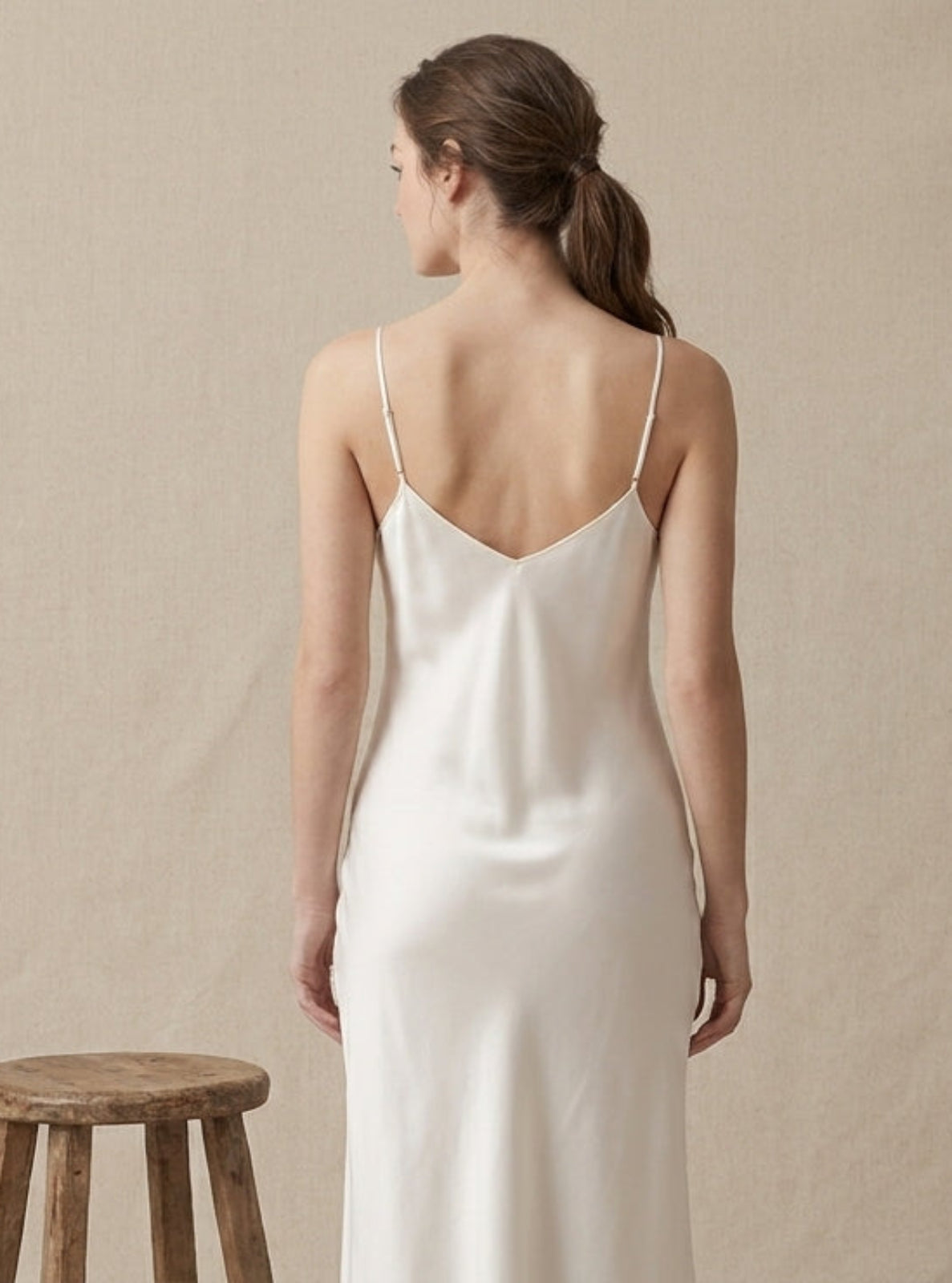 A woman stands with her back to the camera wearing a white satin slip dress with thin spaghetti straps and a low open back, set against a draped beige fabric backdrop with a small wooden stool at her side.