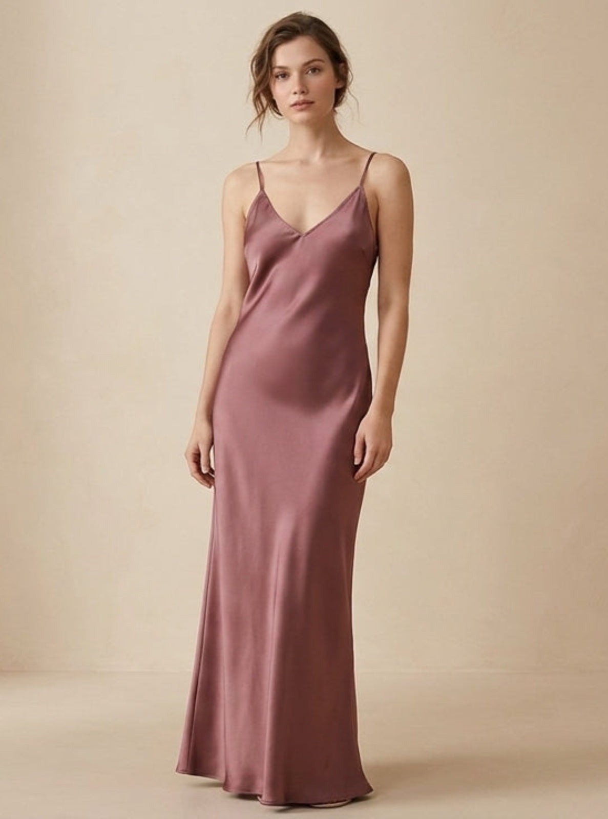 A woman stands in a studio wearing a floor-length satin slip dress in a muted dusty rose or mauve tone against a warm beige backdrop.