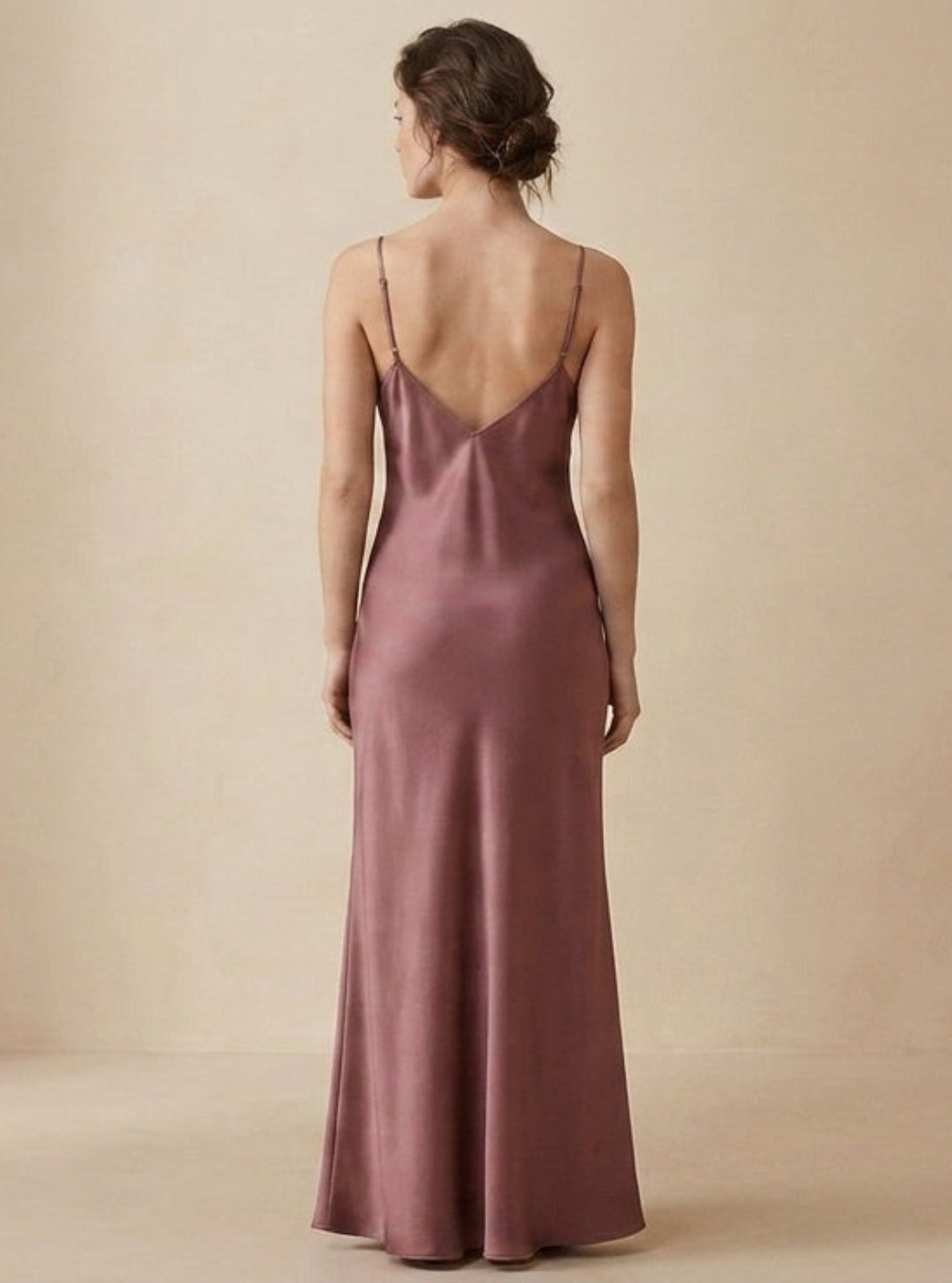 A woman viewed from the back wearing a floor-length satin slip dress in a dusty rose or mauve tone against a plain warm beige studio background.