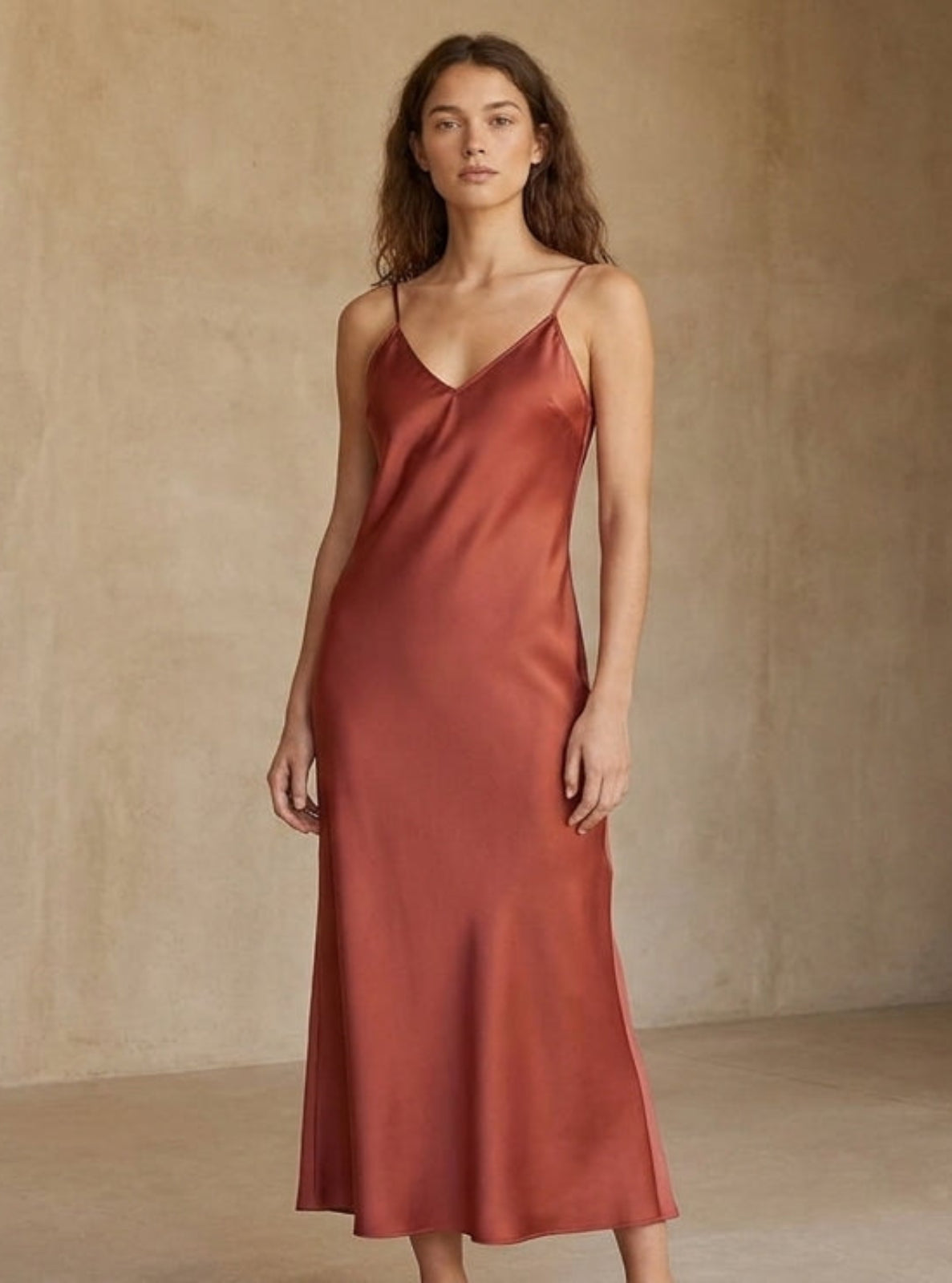 A woman modeling a rust-colored satin slip dress in a minimalist studio with textured beige walls and floor.