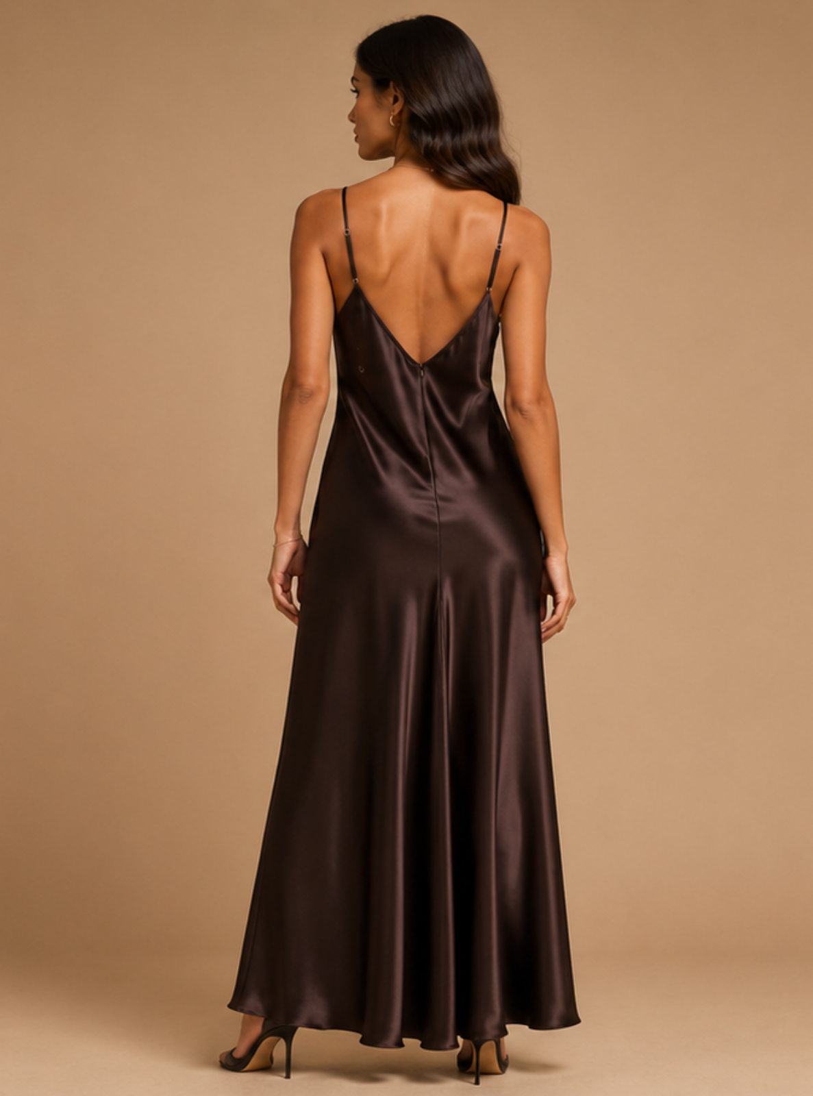 A woman modeling a floor-length satin slip dress in a deep chocolate brown color, standing against a solid beige studio backdrop.