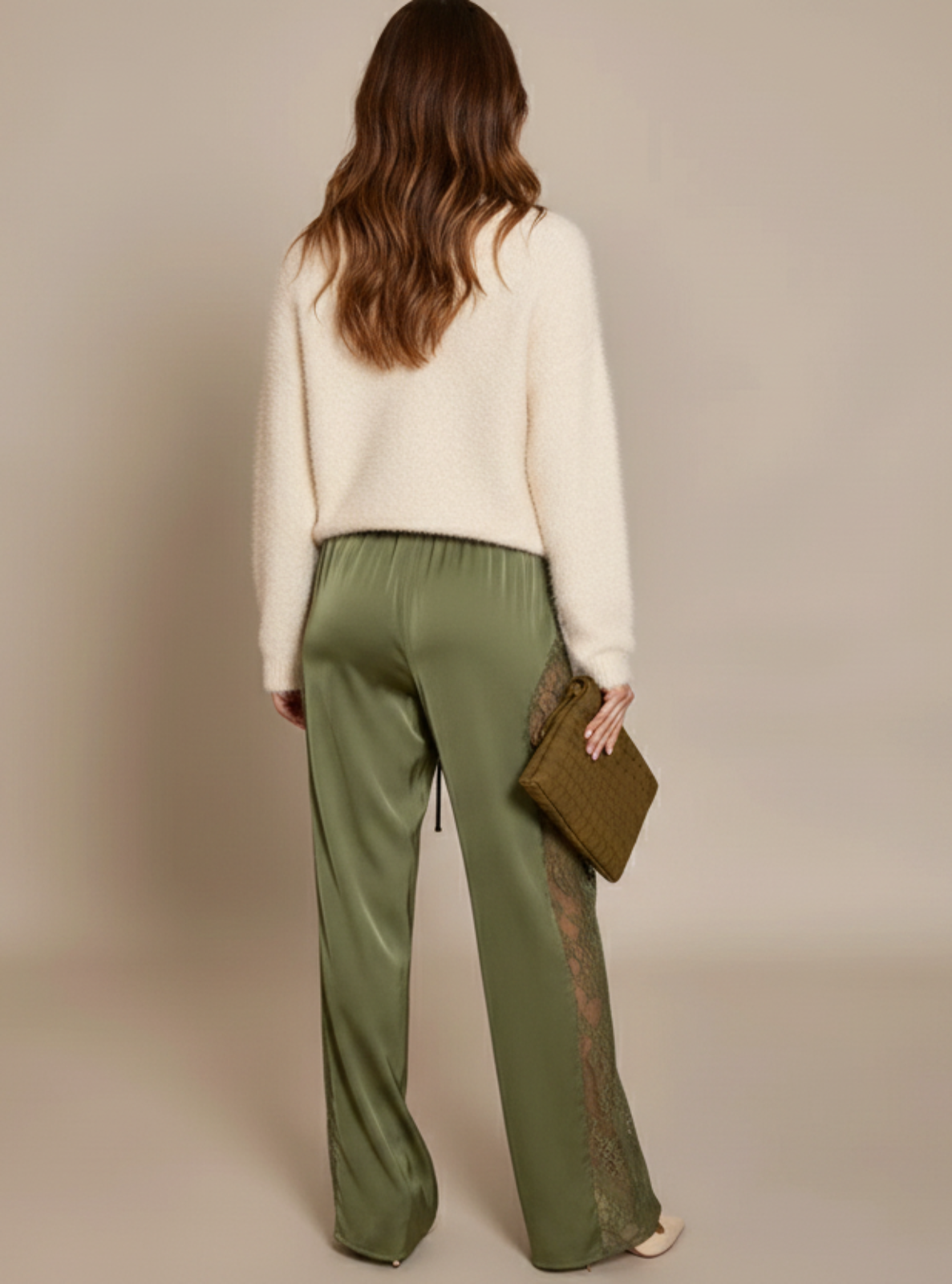 Woman wearing a cream sweater and green pants holding a clutch against a beige background