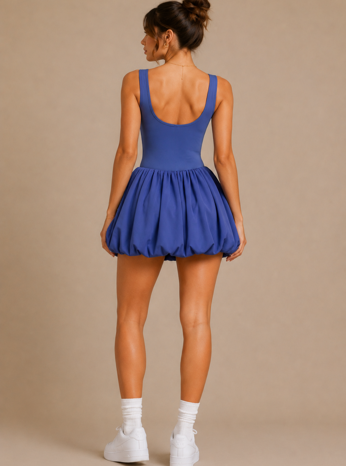 A woman shown from the back wearing a royal blue sleeveless bodysuit with a flared skirt, paired with white socks and white sneakers, against a solid beige studio background.