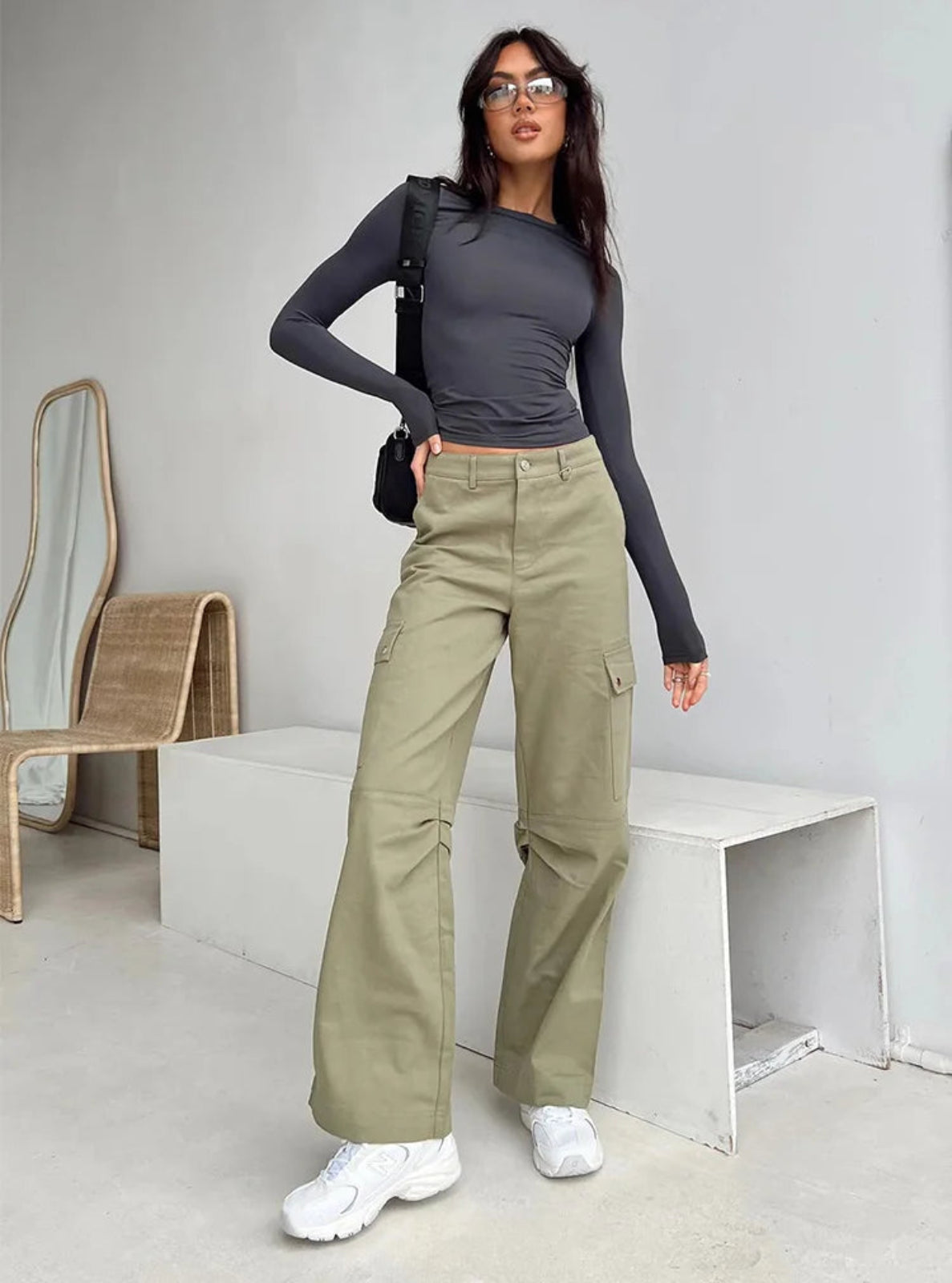 Woman wearing a dark long-sleeve top and light green cargo pants in a minimalistic room.