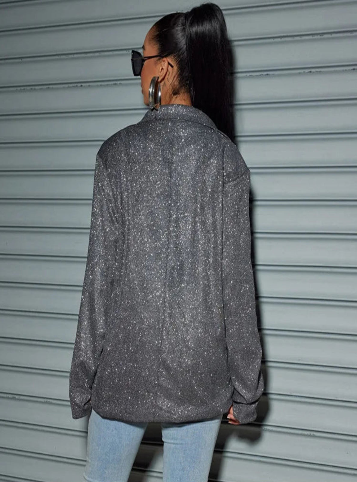 Person wearing a sparkly gray jacket and light blue jeans standing in front of a gray metal shutter.