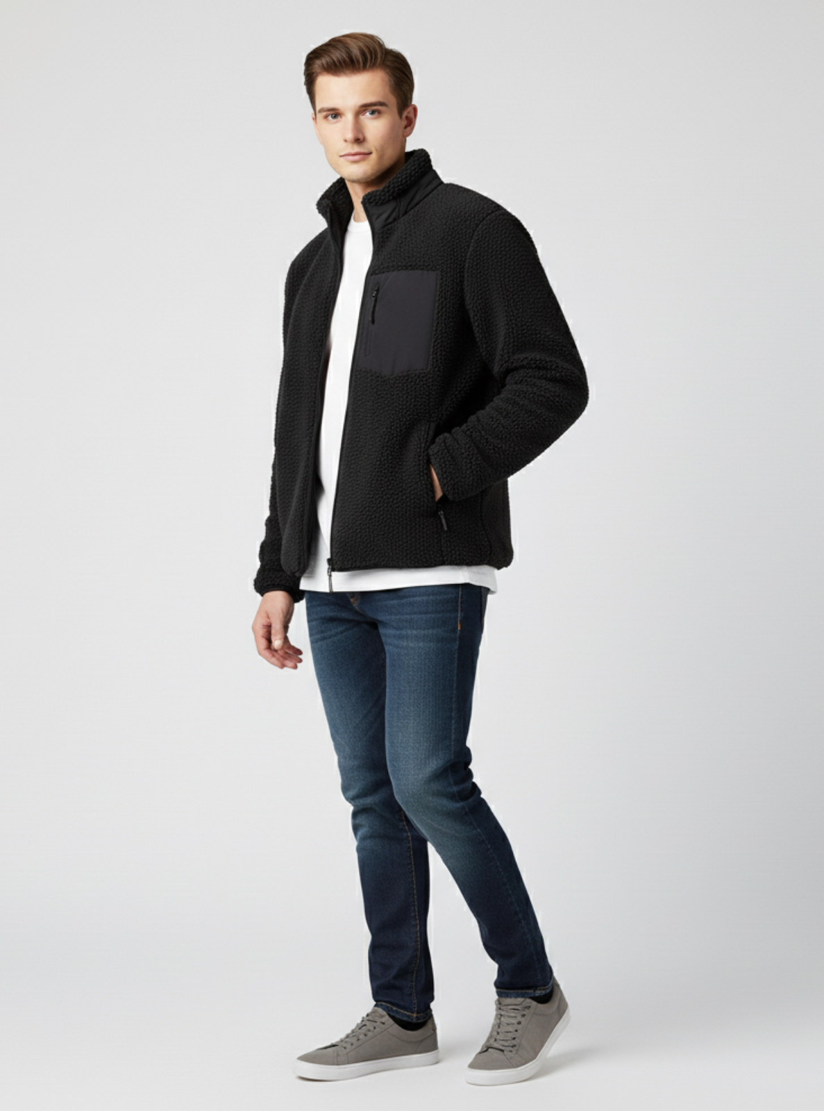 Man wearing a black jacket, white shirt, blue jeans, and gray shoes on a light gray background