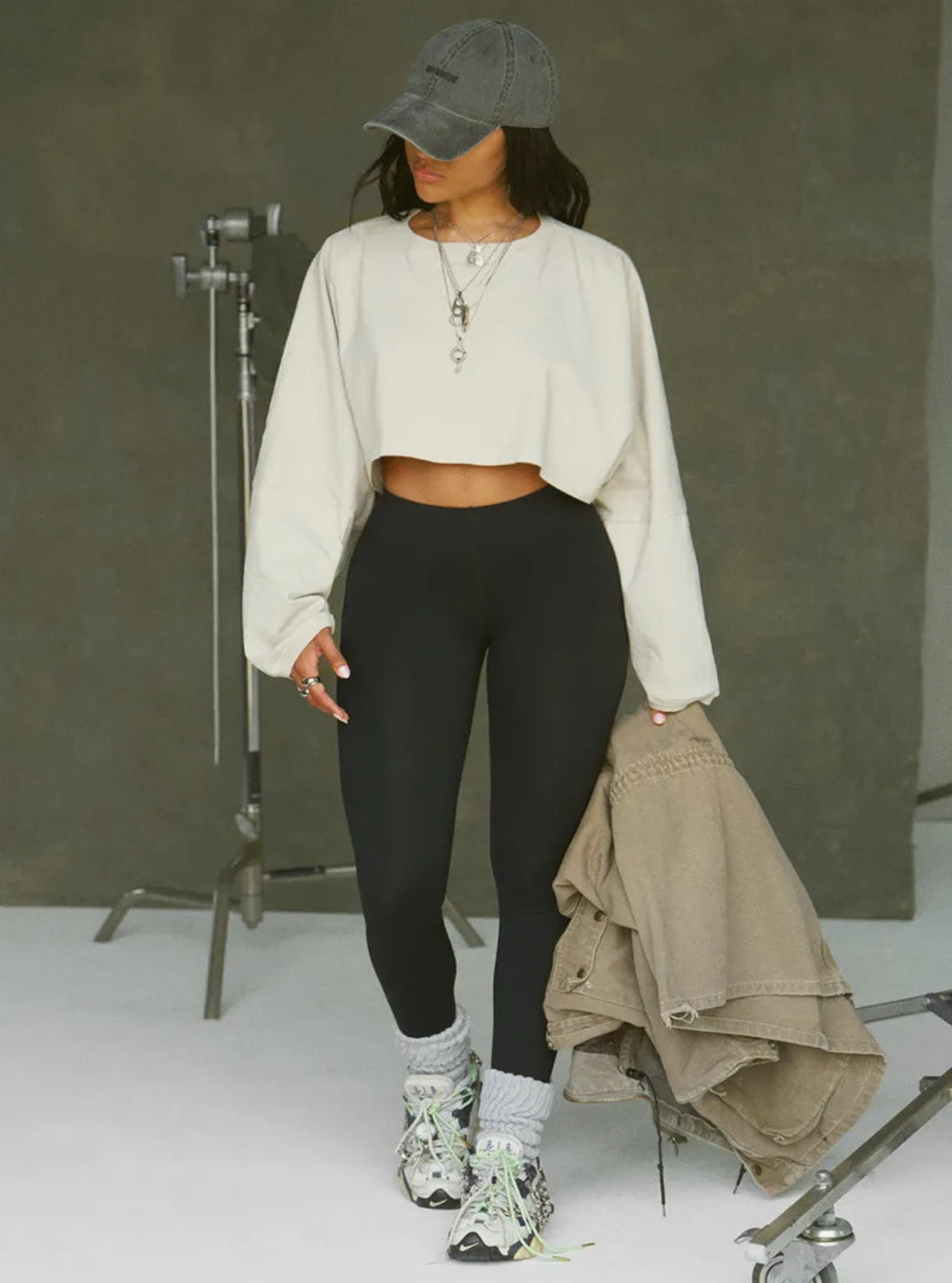Woman wearing a white cropped sweatshirt, black leggings, and a gray cap in a studio setting.