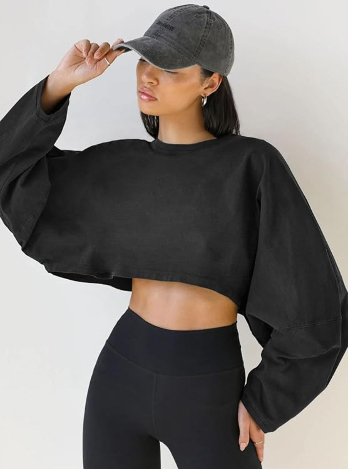 Person wearing a black cropped sweatshirt and leggings with a gray cap on a plain background