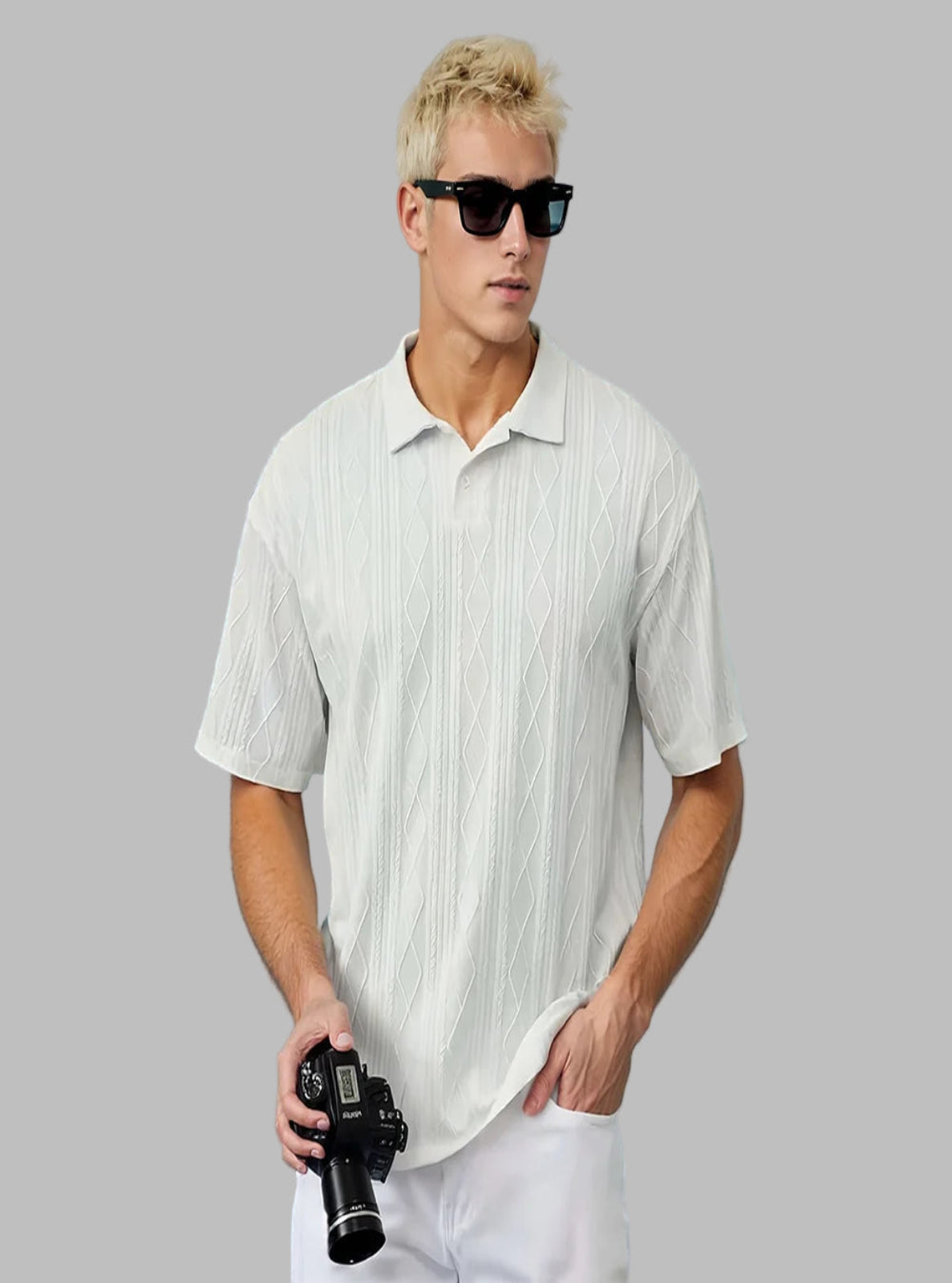 Man wearing a white textured polo shirt and sunglasses, holding a camera against a light blue wall.