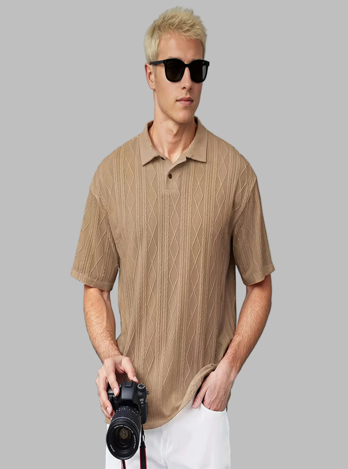 Man wearing a beige textured polo shirt and sunglasses, holding a camera against a light blue wall.