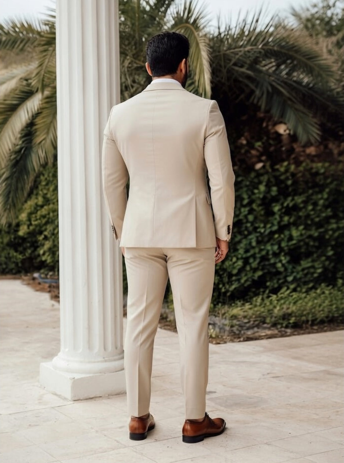 Man in a beige suit standing in front of classical columns with greenery in the background