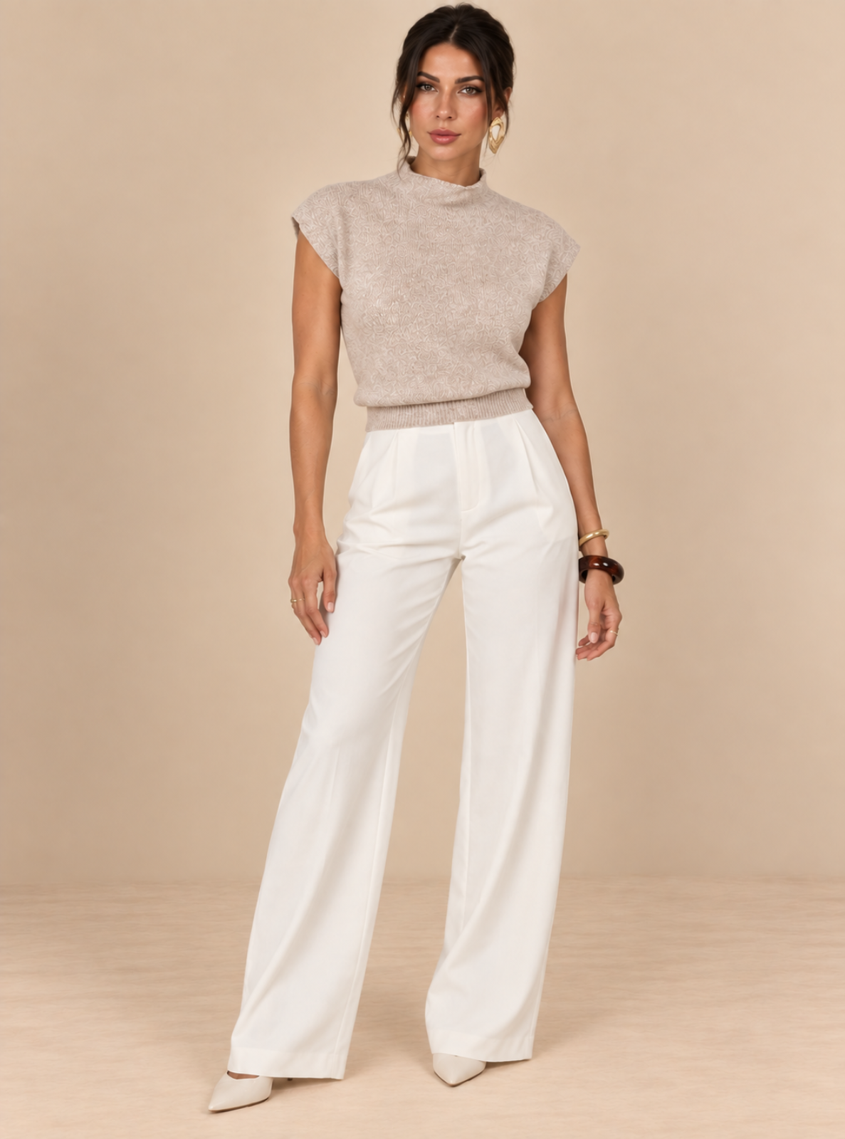 A woman modeling a light beige textured knit mock-neck top paired with white wide-leg trousers, standing against a solid beige studio backdrop.