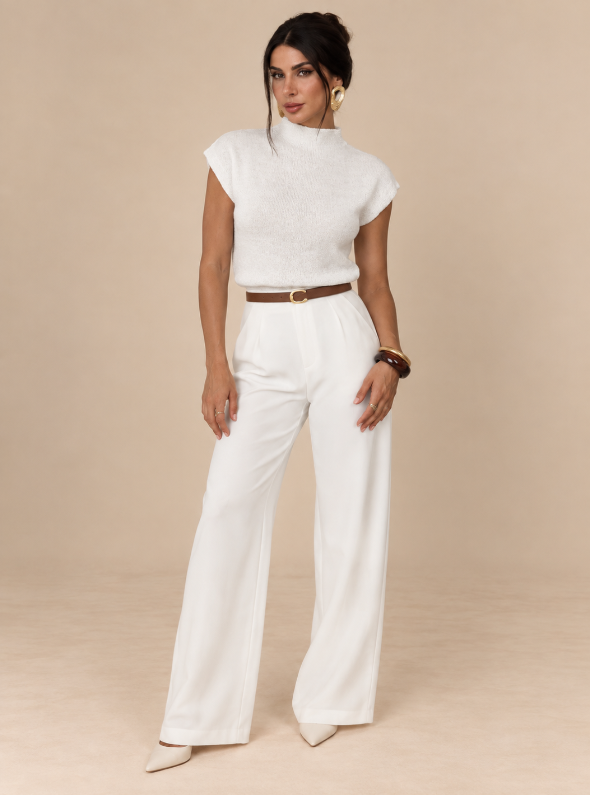 A woman modeling a white textured short-sleeve mock-neck top and matching white wide-leg trousers with a brown belt, standing against a solid beige studio background.