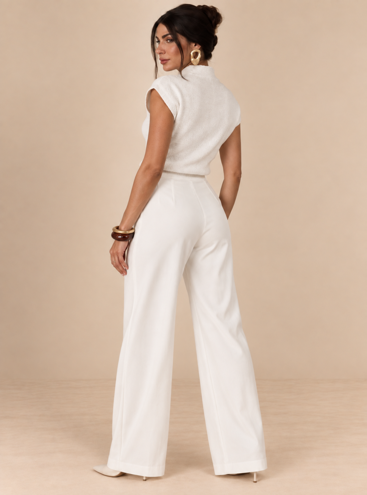 A woman modeling a white sleeveless jumpsuit from the back, standing against a solid beige studio background.