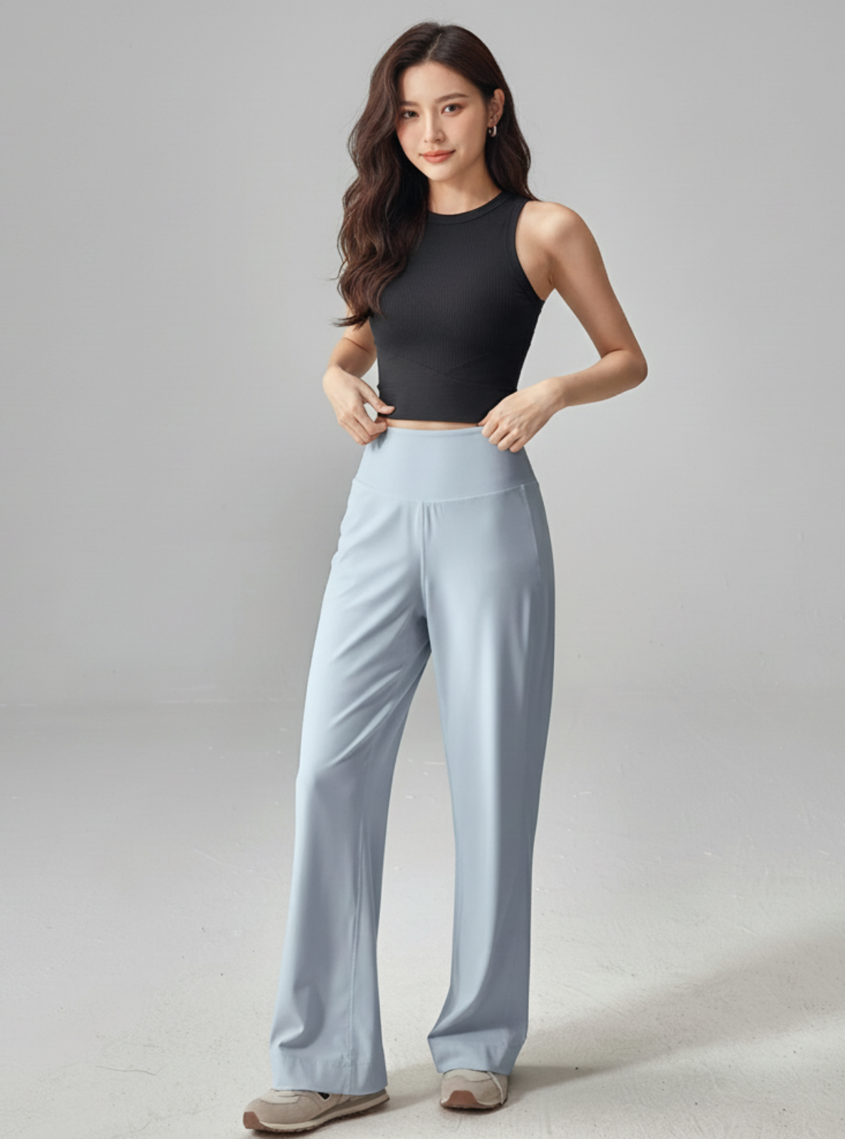 Woman wearing a black sleeveless top and light blue pants on a gray background