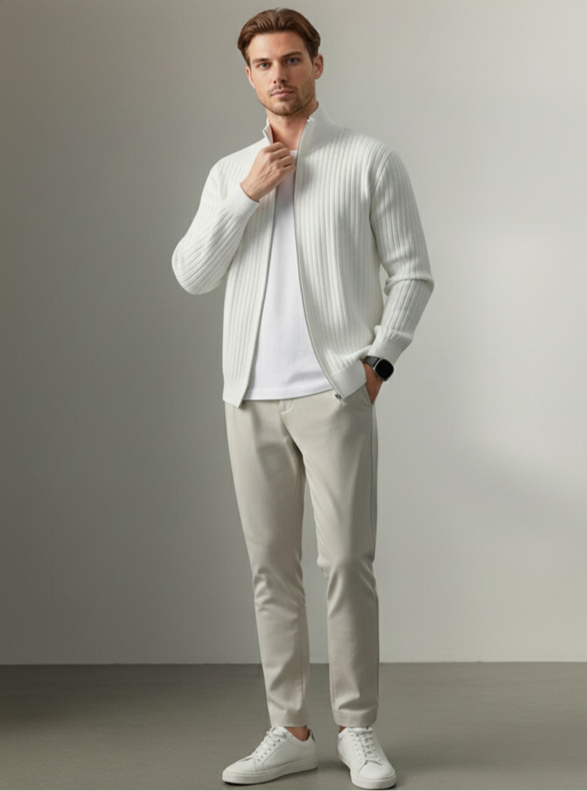 Man wearing a white ribbed sweater, white shirt, beige pants, and white sneakers against a plain background