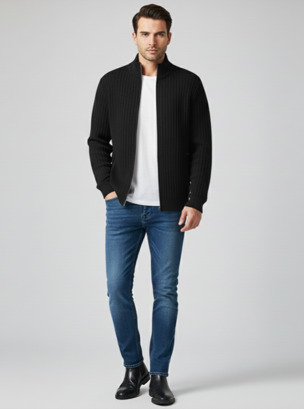 Man wearing a black cardigan, white shirt, blue jeans, and black shoes on a gray background