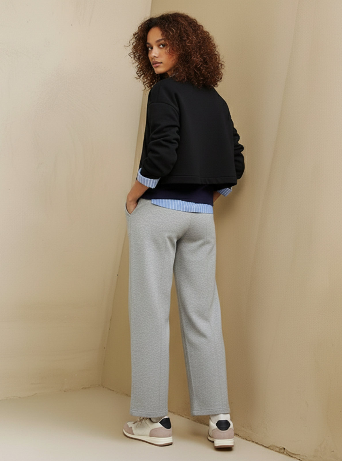 Woman wearing a black jacket and gray pants against a beige wall