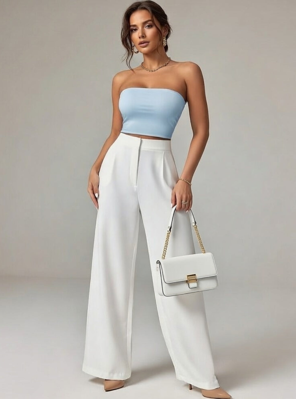 Woman wearing a light blue strapless top and white wide-leg pants, holding a white handbag against a neutral background.