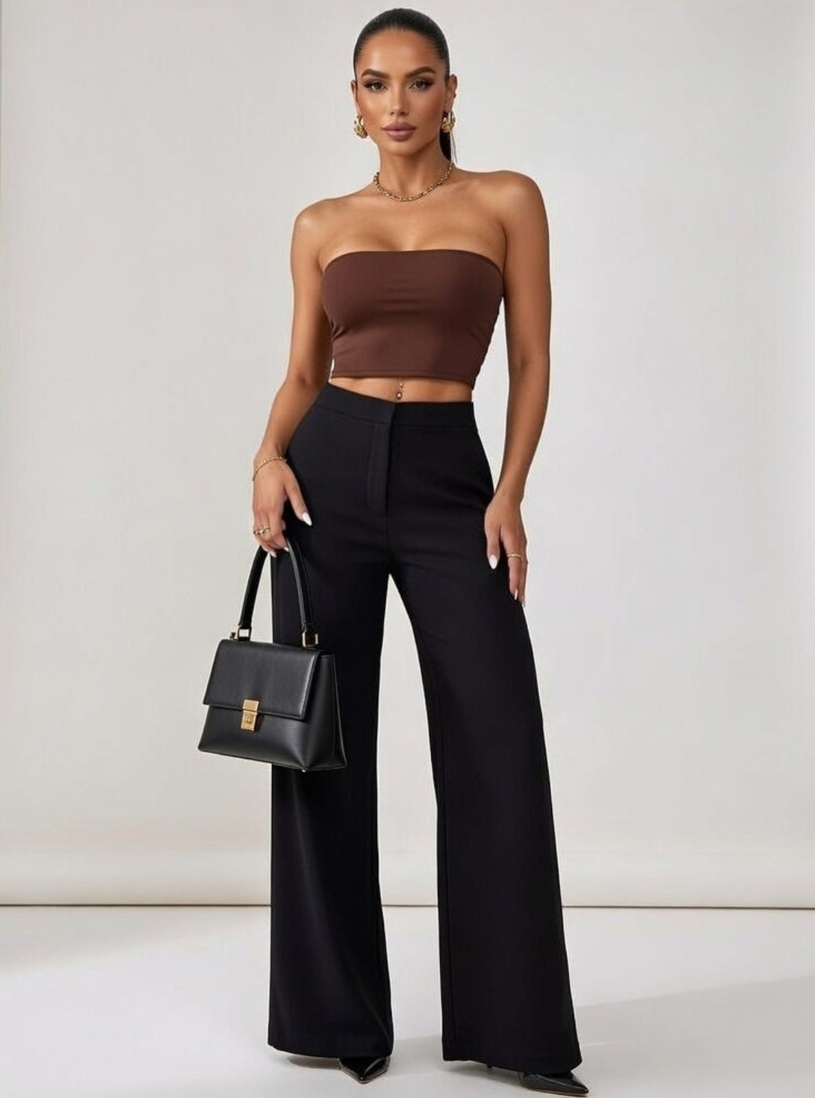 Woman wearing a brown strapless top and black high-waisted pants, holding a black handbag on a plain background.